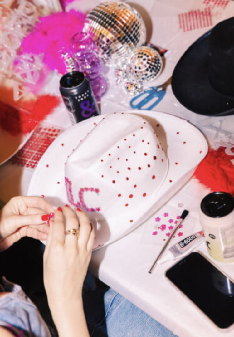 A person decorating a white hat with red polka dots using pink and purple embellishments at a party or craft event. The table has various decorations, accessories, and a drink in a cup.