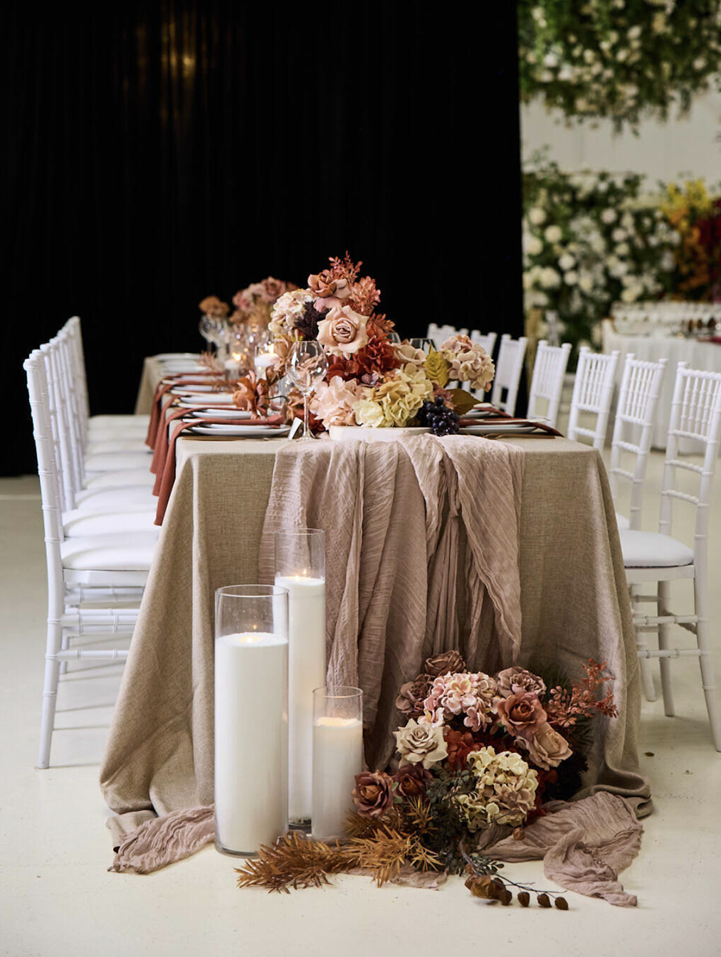 An elegantly decorated banquet table with floral centerpieces in soft pink, cream, and mauve tones, surrounded by white chairs, candles, and draped fabric, set for a wedding or special event.