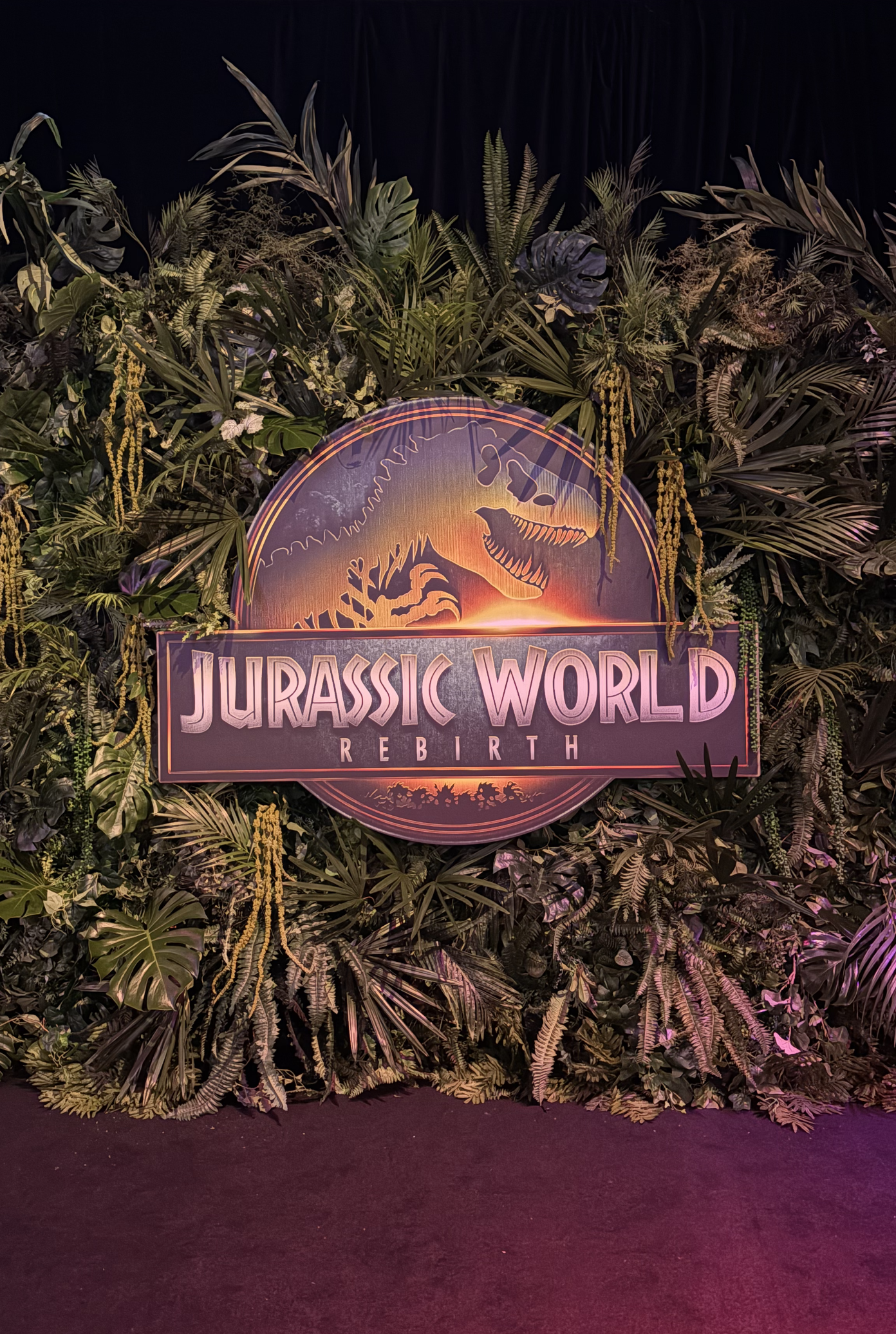 A jungle-themed backdrop with lush green tropical leaves and plants surrounding a circular sign that reads 'Jurassic World Rebirth' with an image of a dinosaur.