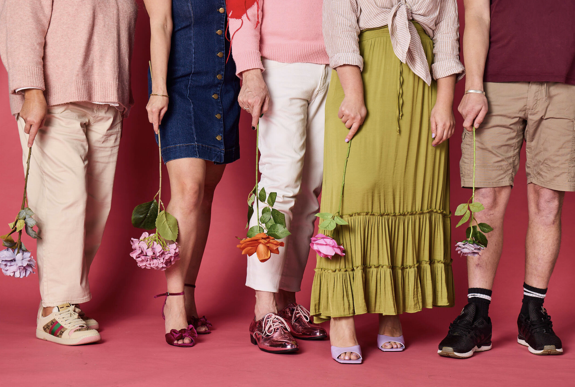 Six people standing against a pink background, holding colorful flowers. The image shows their lower bodies, dressed in a variety of clothing styles, including pants, skirts, and shorts, with different types of footwear.