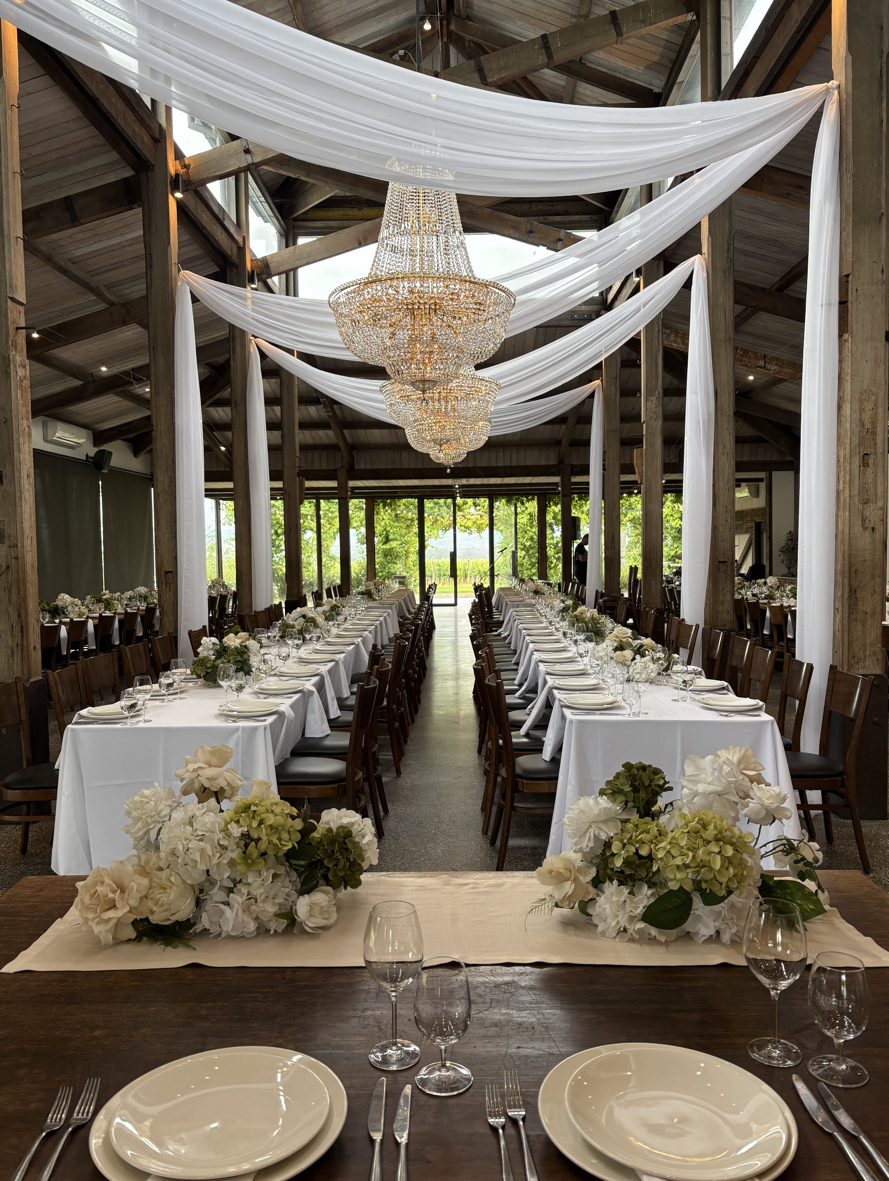Elegant wedding reception setup with long tables covered with white tablecloths, floral centerpieces, and place settings. The venue has wooden beams, hanging chandeliers, and white drapery decorating the ceiling, with large windows showing greenery o