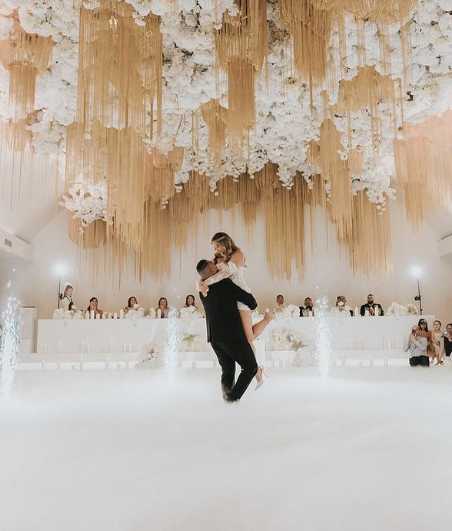 Couple dancing at a wedding reception with floral ceiling decorations and seated guests in the background.