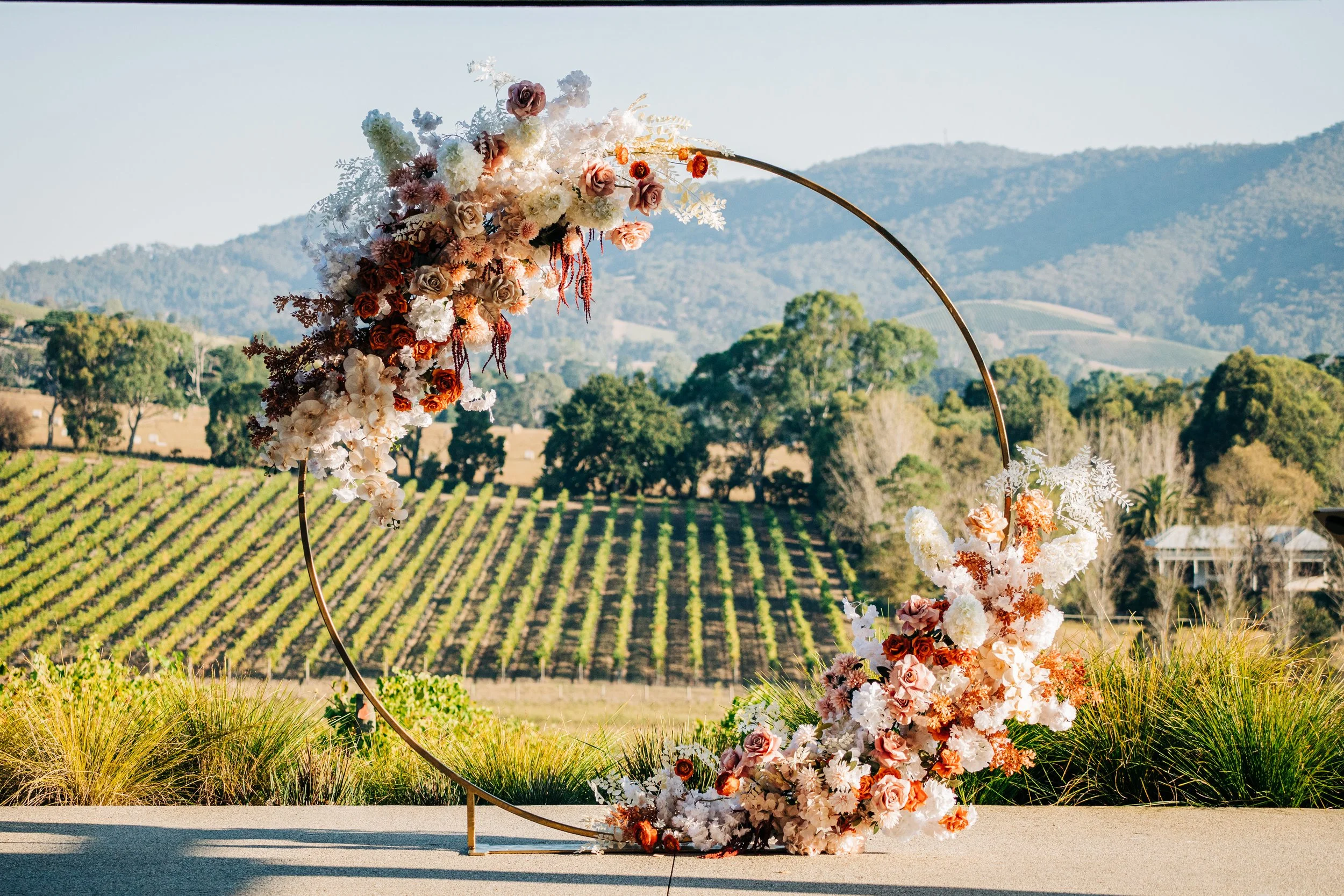 Floral wedding arch with pink, white, and red flowers set outdoors with vineyard and hills in the background.