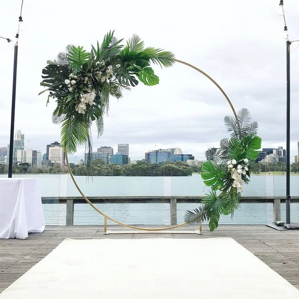 Decorative circular floral arch with tropical green leaves and white flowers on a waterfront deck with city skyline in the background.