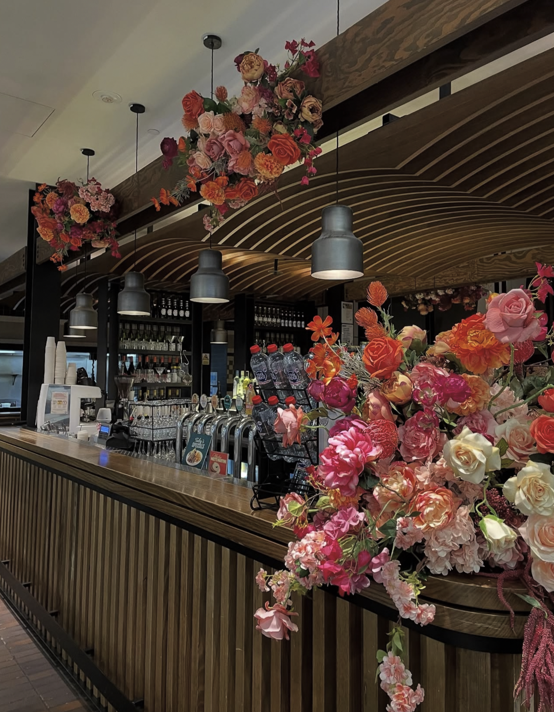 A bar with colorful floral arrangements hanging from the ceiling and a bouquet on the counter, with glasses, bottles, and coffee machine behind the bar.