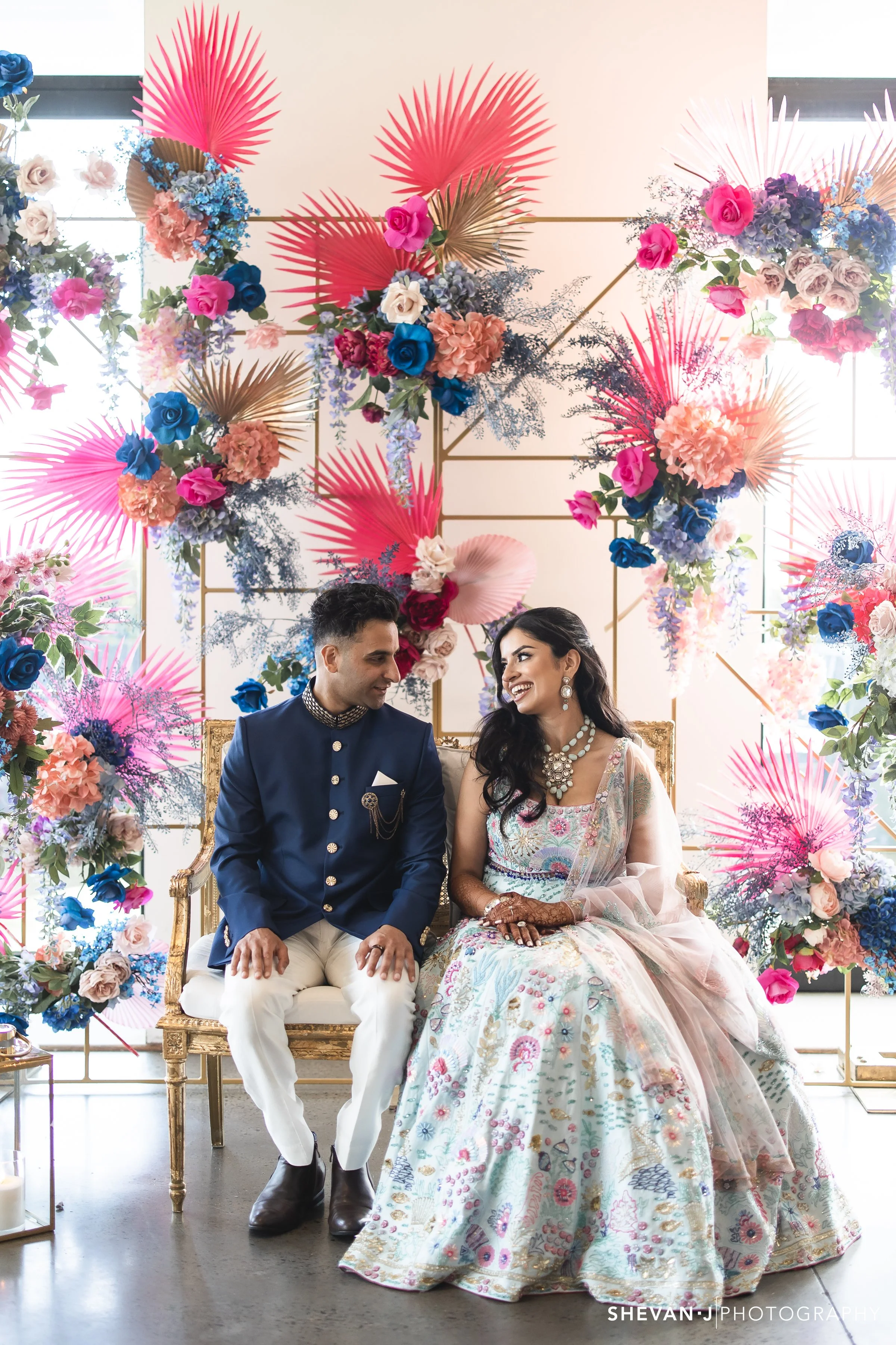 A couple dressed in formal South Asian attire sitting on a gold ornate sofa, surrounded by colorful floral decorations including pink, blue, and purple flowers with pink and gold palm leaves, set against a bright backdrop with large windows.