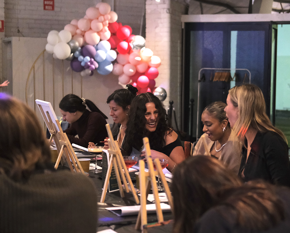 Group of women sitting at a table, laughing and socializing at a celebration or event with balloon decorations in the background.