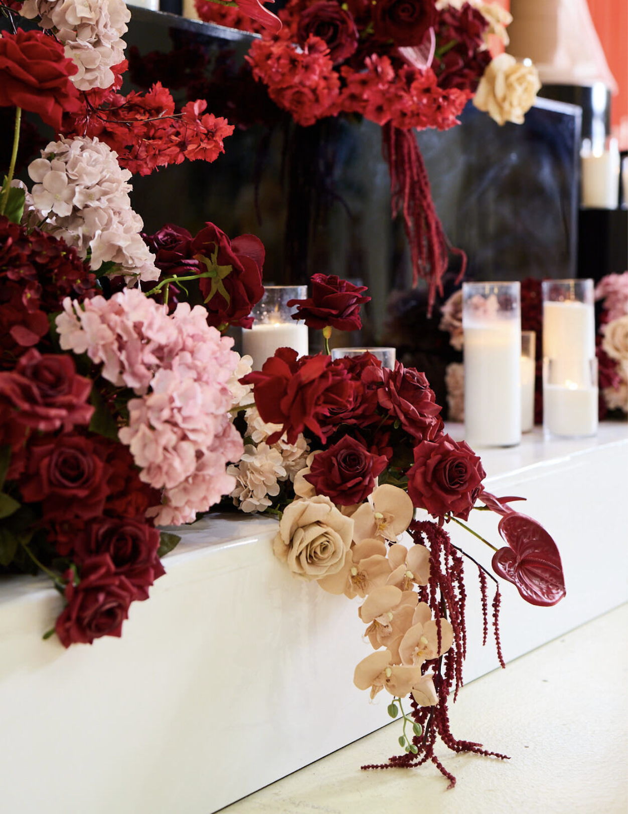 A floral arrangement with red, pink, and beige roses, pink hydrangeas, and red and beige orchids, decorated with candles in glass holders, placed on a white surface in front of a dark backdrop with candles.