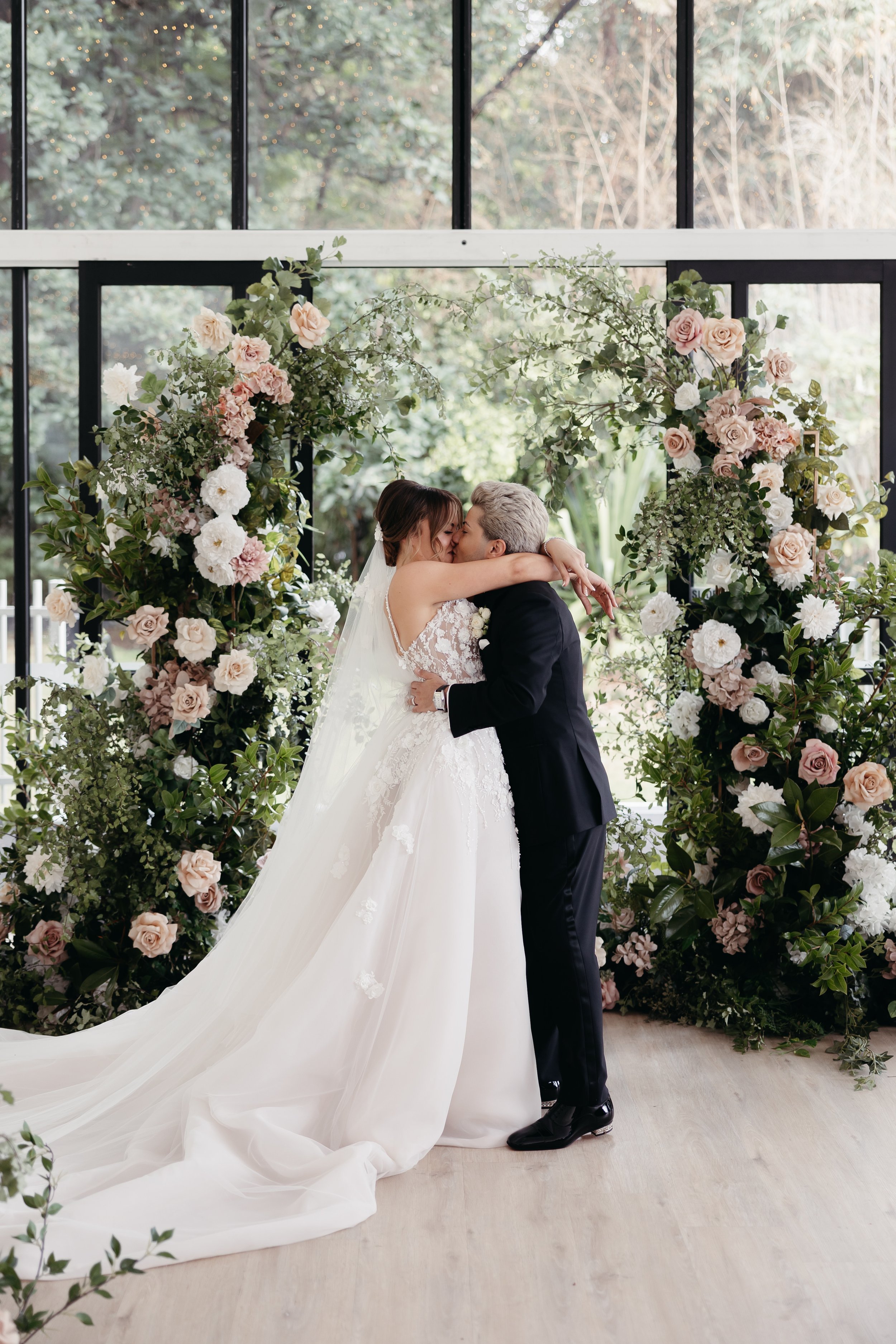 A bride and groom sharing a kiss at their wedding ceremony, standing in front of a floral arch with white, pink, and green flowers, indoors with large windows and fairy lights outside.