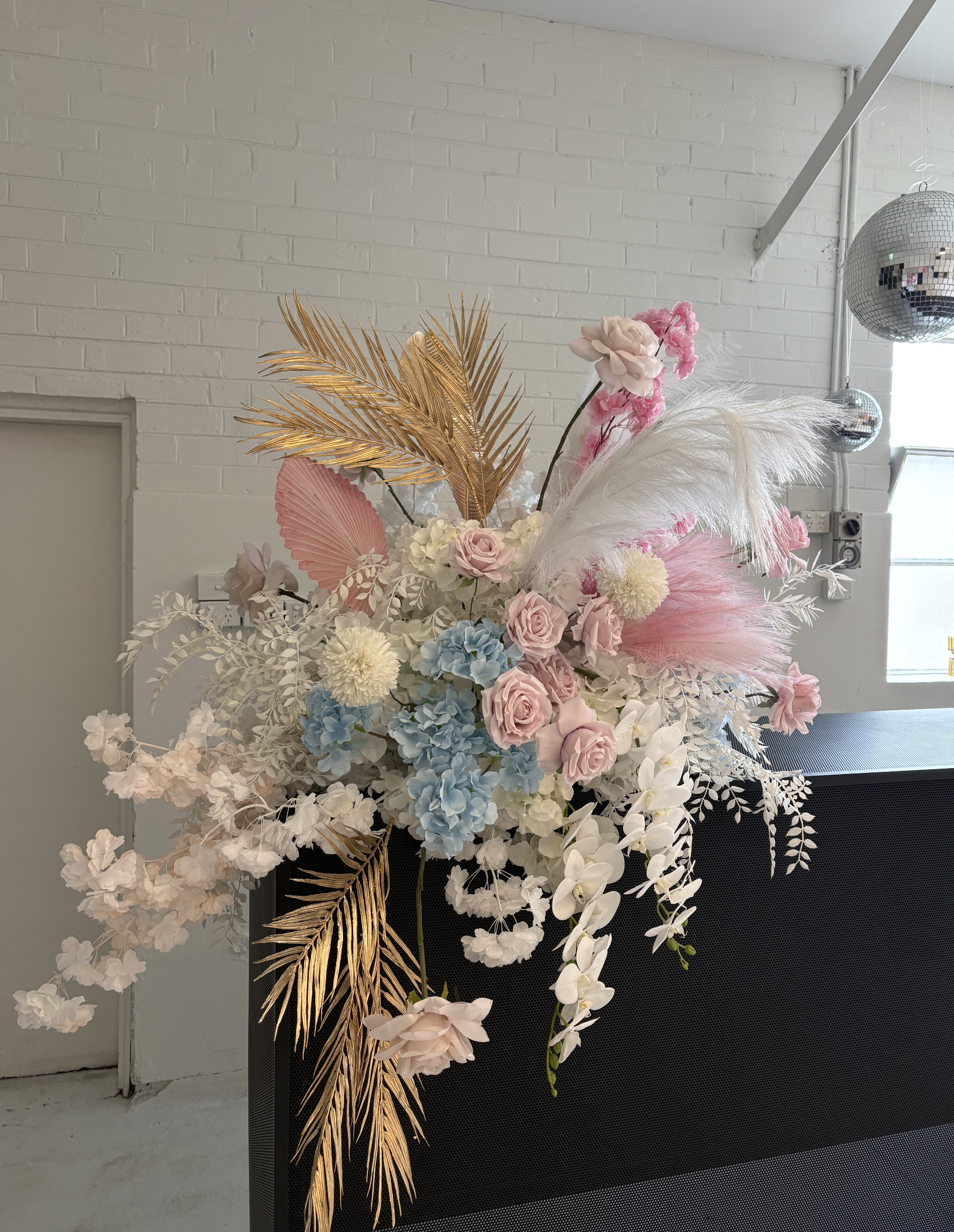 A large floral arrangement with pink, white, and blue flowers, including roses, orchids, and hydrangeas, accented with gold-painted palm leaves, pink feathers, white pampas grass, and pink palm leaves, placed on a black surface against a white brick 