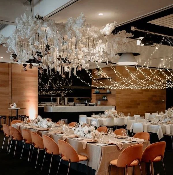 Decorated banquet hall with hanging white floral arrangements, string lights, and tables set with white tablecloths, floral centerpieces, and place settings for a special event.