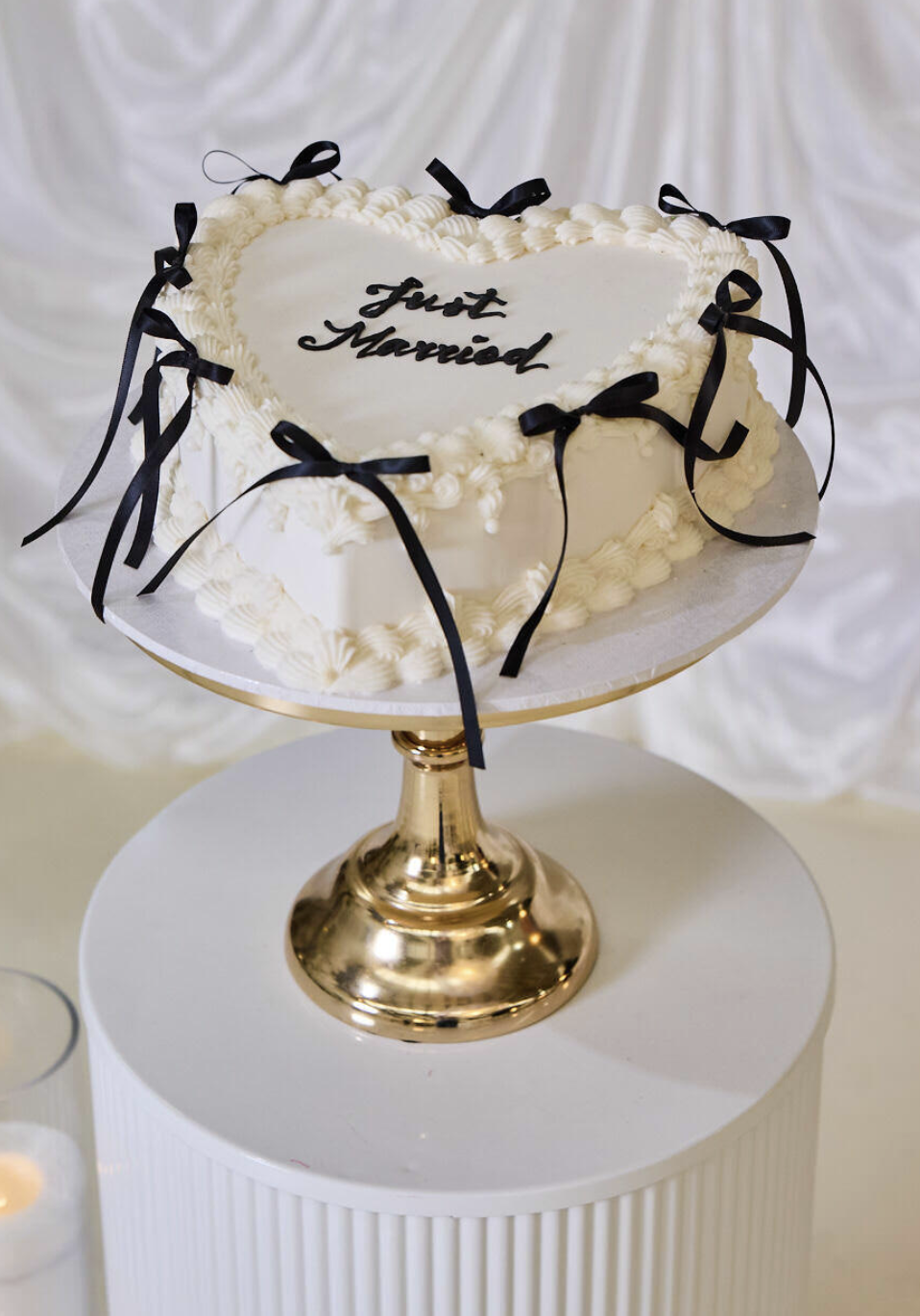 White heart-shaped wedding cake with black ribbons, decorative frosting, and 'Just Married' written on top, displayed on a gold cake stand.