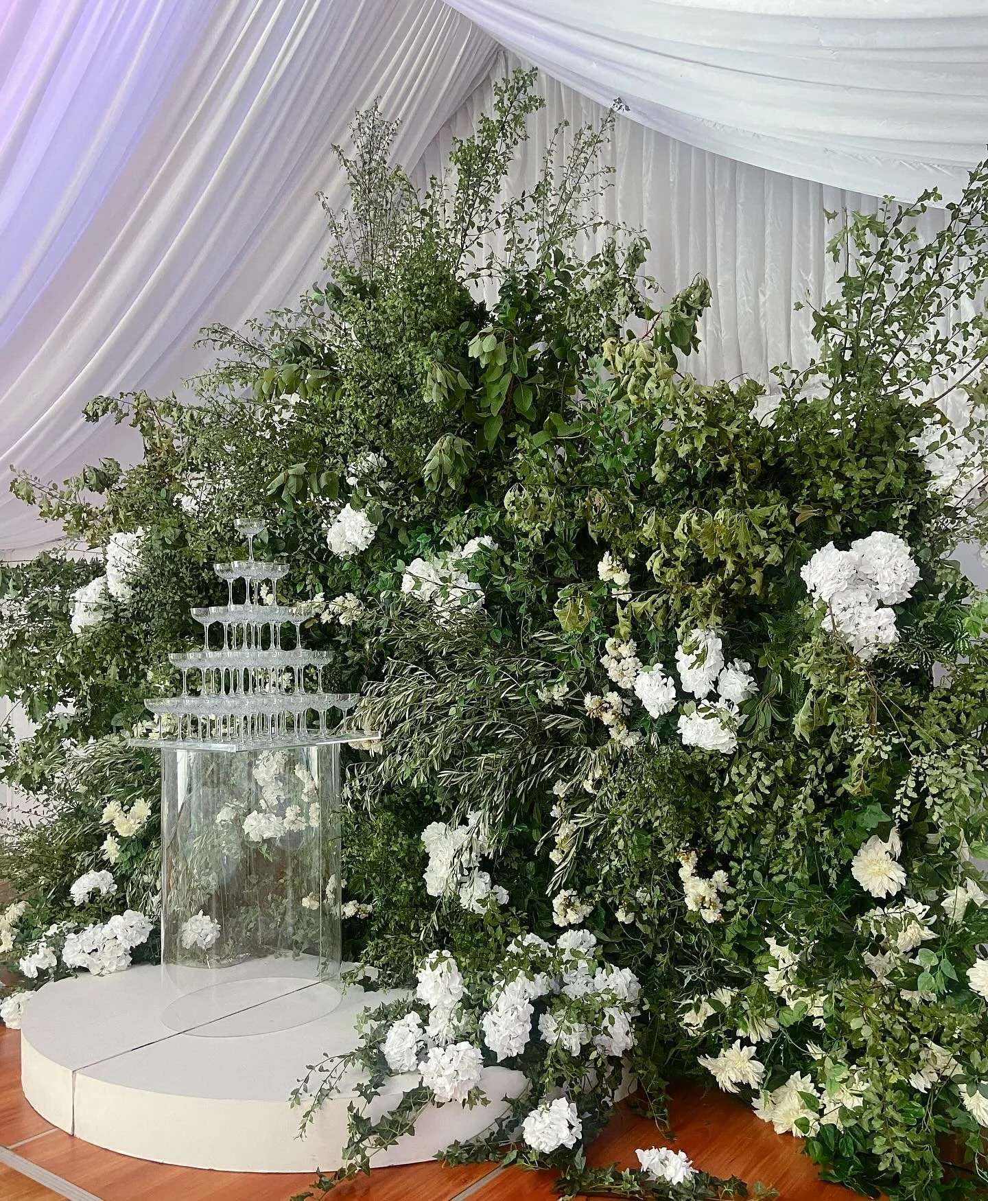 A floral arrangement featuring white flowers and green foliage on a white circular platform, with a champagne glass tower centerpiece, set against a backdrop of white draped fabric.