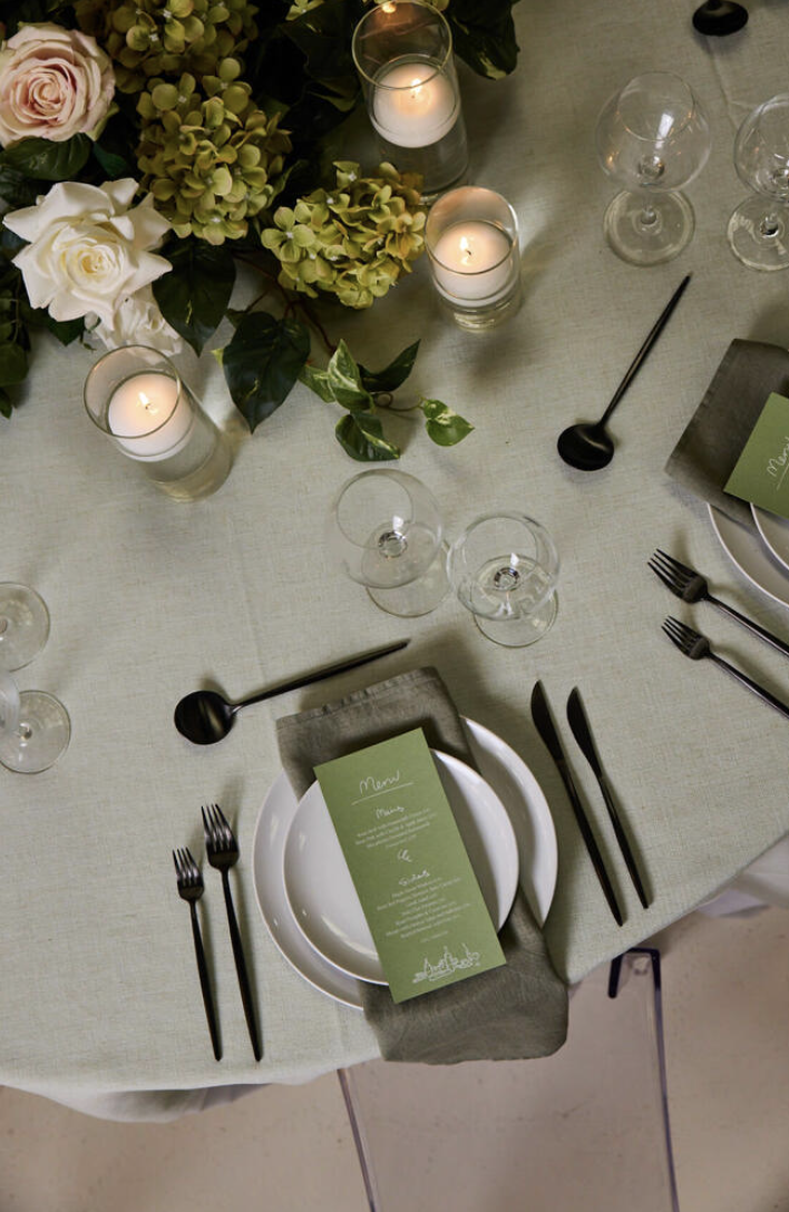 A table setting with plates, black utensils, floral centerpiece with pink roses and green hydrangeas, candles in glass holders, empty wine glasses, and a green menu card.