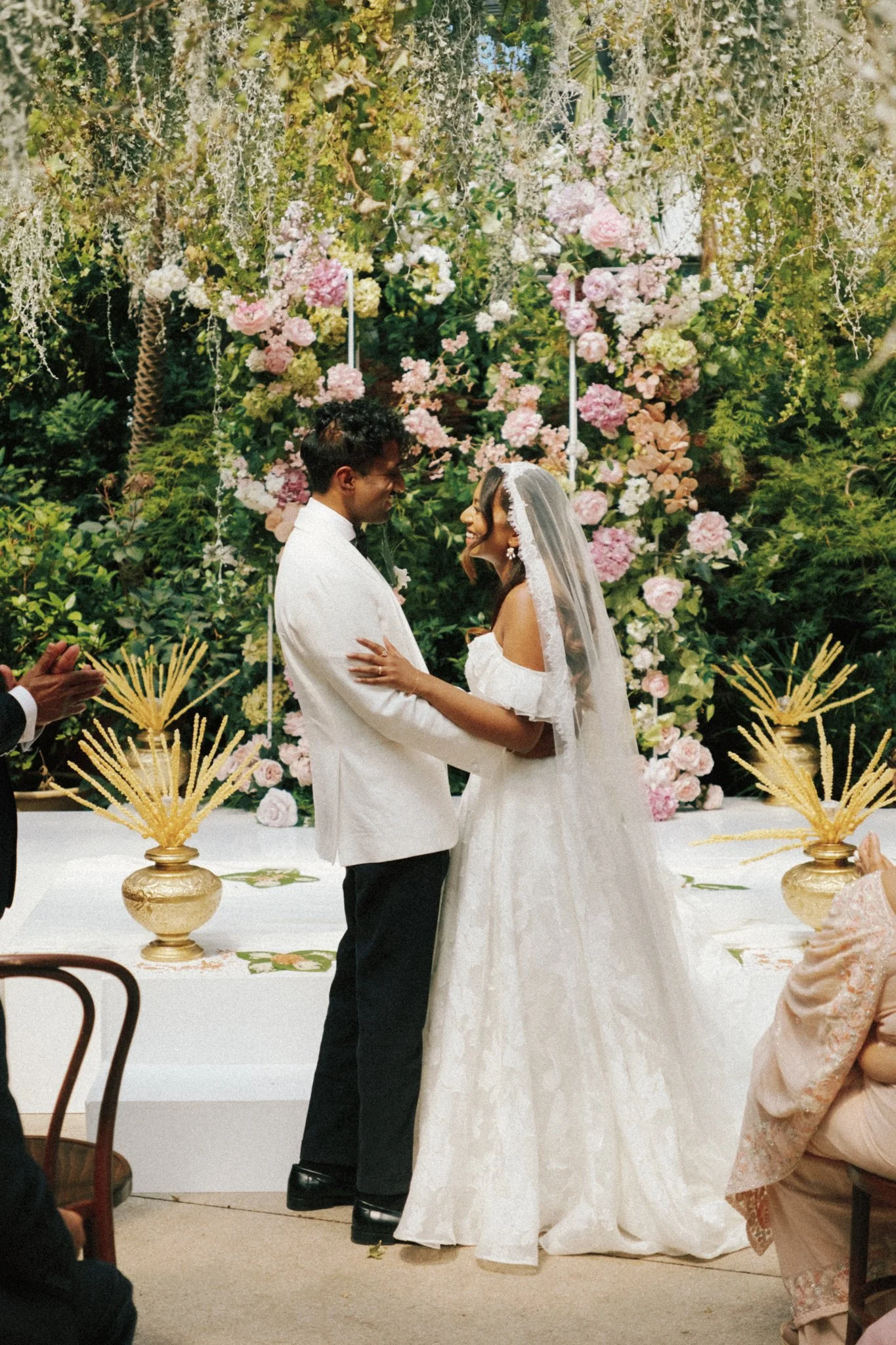 A bride and groom exchanging vows during their wedding ceremony, standing in front of a floral backdrop with pink and white flowers, surrounded by guests.
