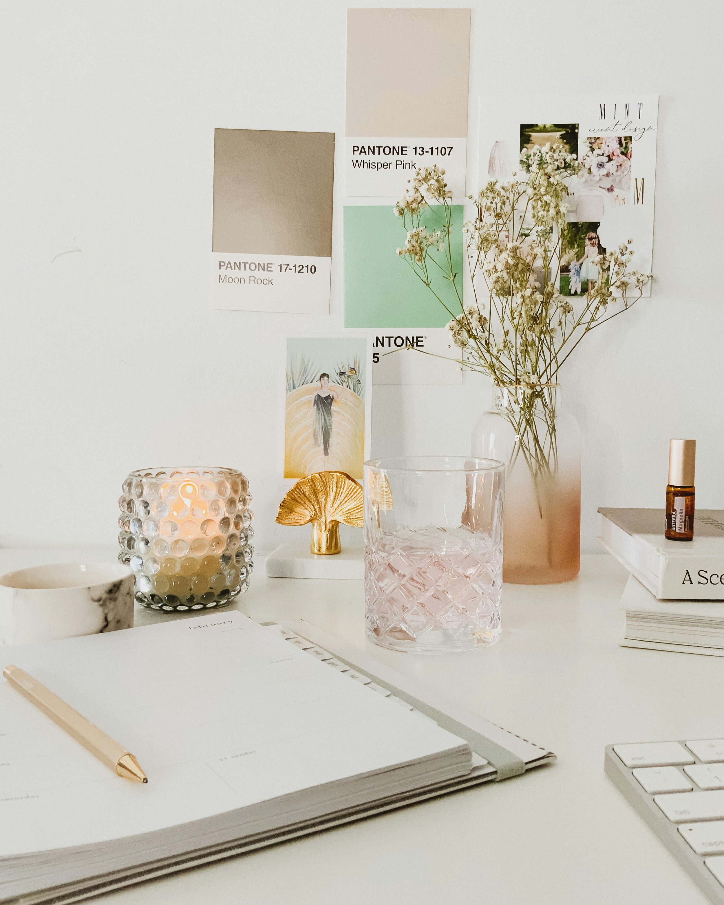 A white desk with a notebook, a gold pen, a marble-patterned bowl, a textured candle holder, a small white dish, a gold mushroom decor piece, a glass of water, a pink vase with dried flowers, a stack of books, a small essential oil bottle, and a computer keyboard. Behind the desk, there is a white wall with color swatches, photos, and a postcard.