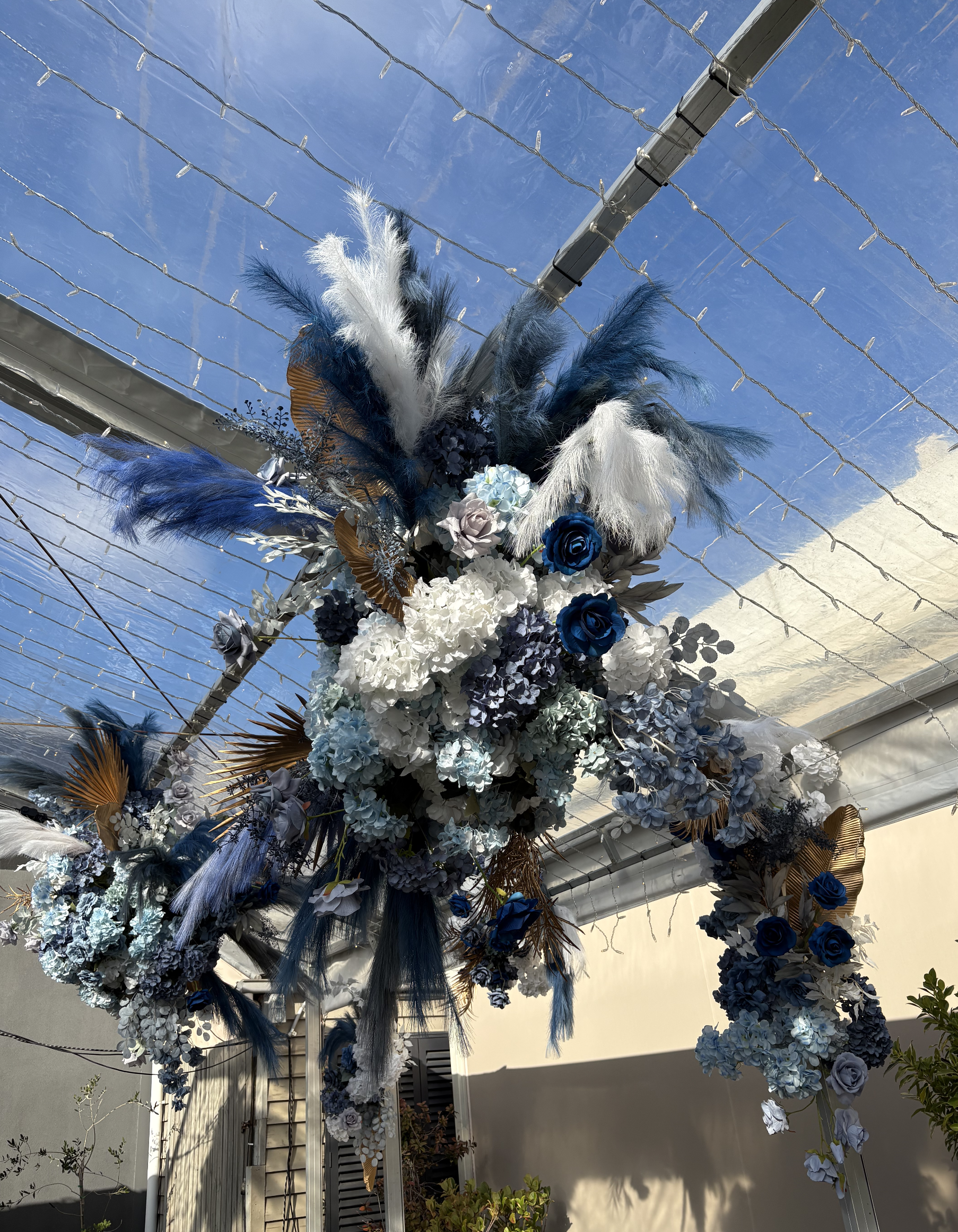 Floral arch with white and blue flowers, feathers, and gold accents under a clear plastic canopy and string lights.