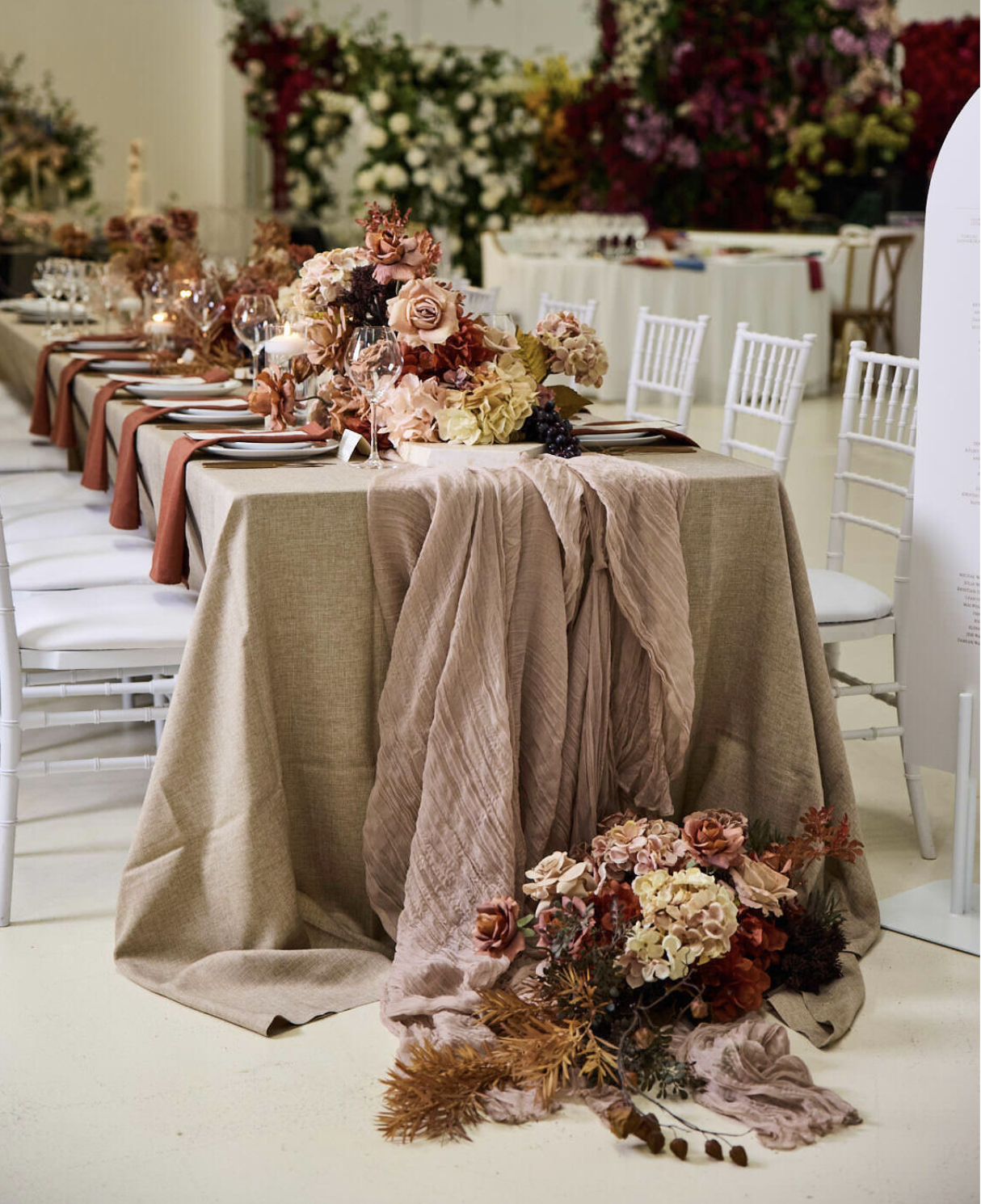 Elegant wedding table setting with a long beige tablecloth, floral centerpiece with blush and dark flowers, wine glasses, and napkins, surrounded by white chairs, in a decorated room with many flowers in the background.