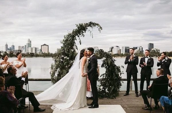 Bride and groom kissing during their outdoor wedding ceremony on a wooden deck by a river with a city skyline in the background. Bridesmaids and groomsmen are clapping.