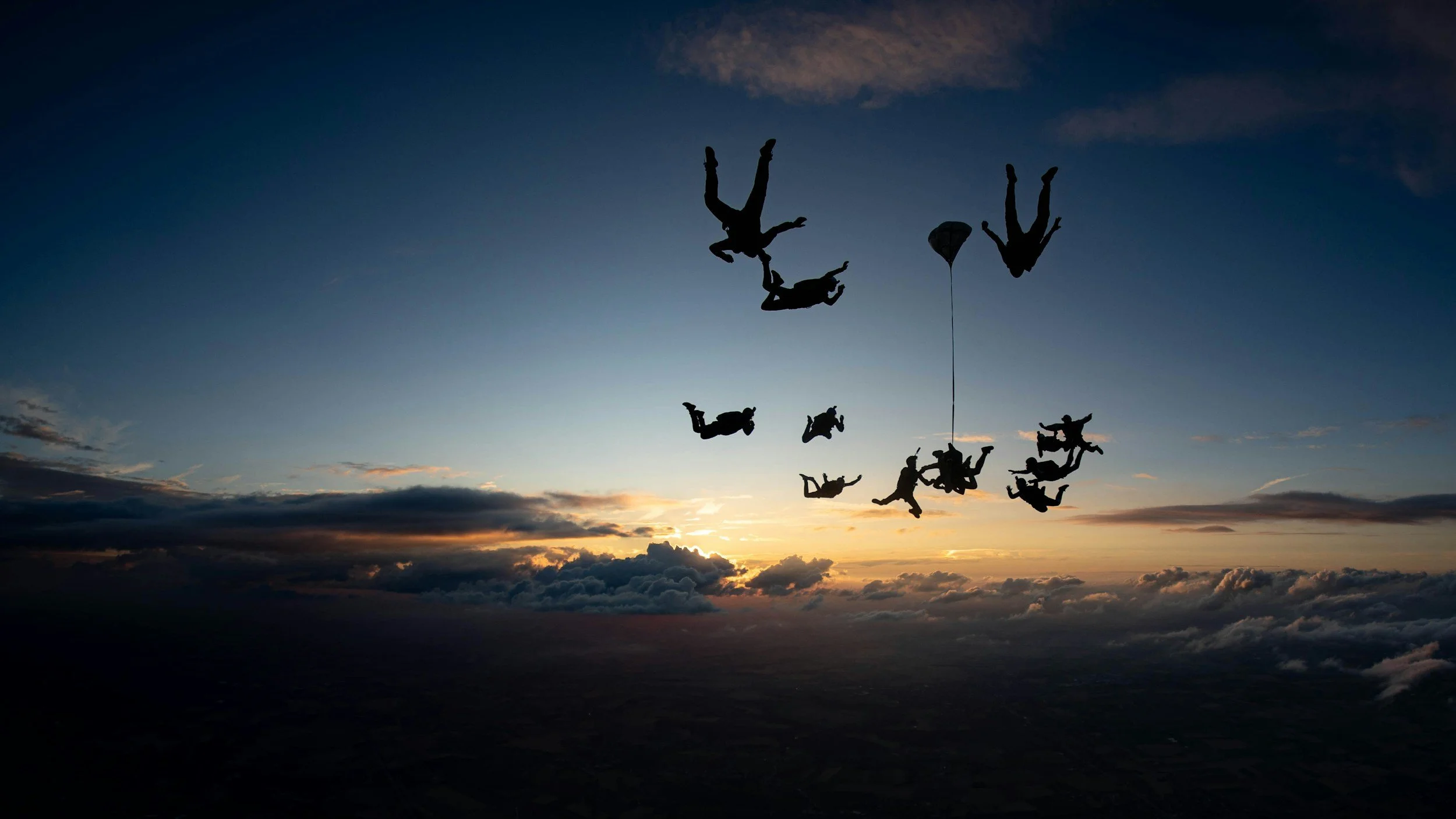 Silhouettes of skydivers holding hands and falling in formation against a sunset sky with clouds.