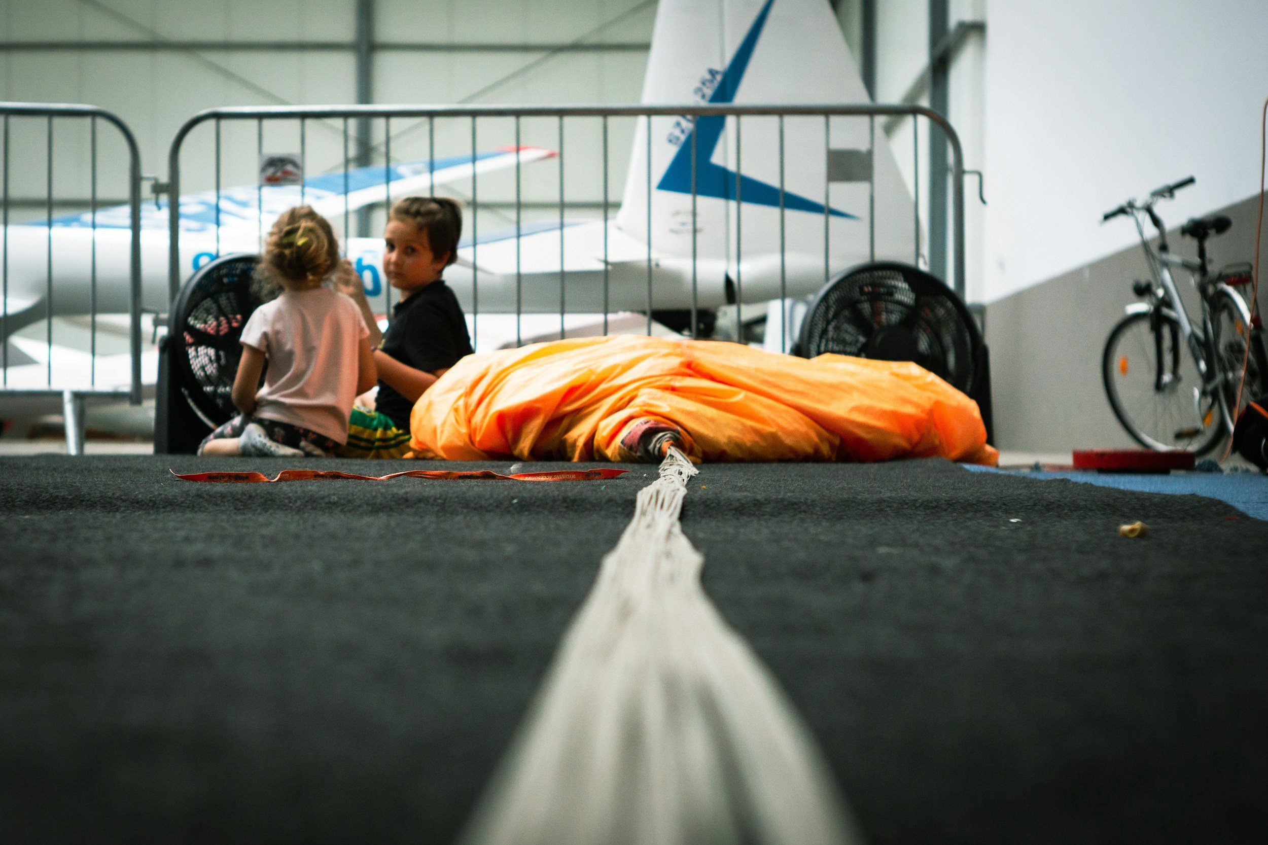 Two children sitting and talking next to a sleeping bag on the floor of an indoor space, with an airplane and bicycles in the background.