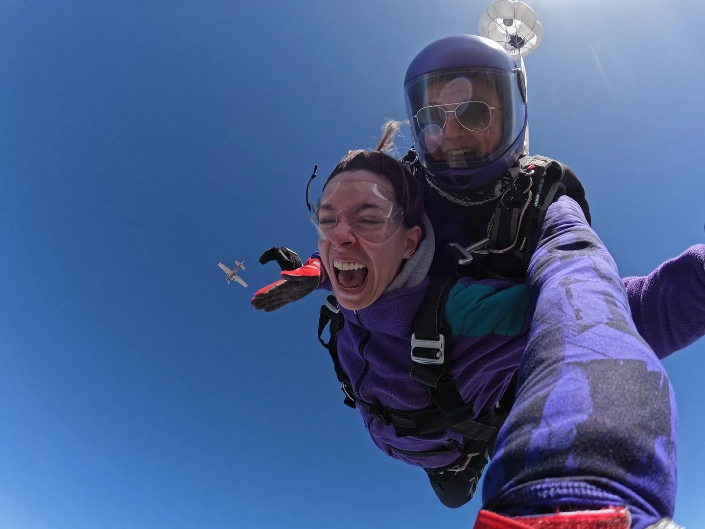Two individuals are skydiving, with one person attached to the other. The person in front is smiling and laughing, wearing glasses and a purple jacket, while the person behind, wearing a helmet and sunglasses, appears to be the instructor. A small airplane is visible in the background against a clear blue sky.