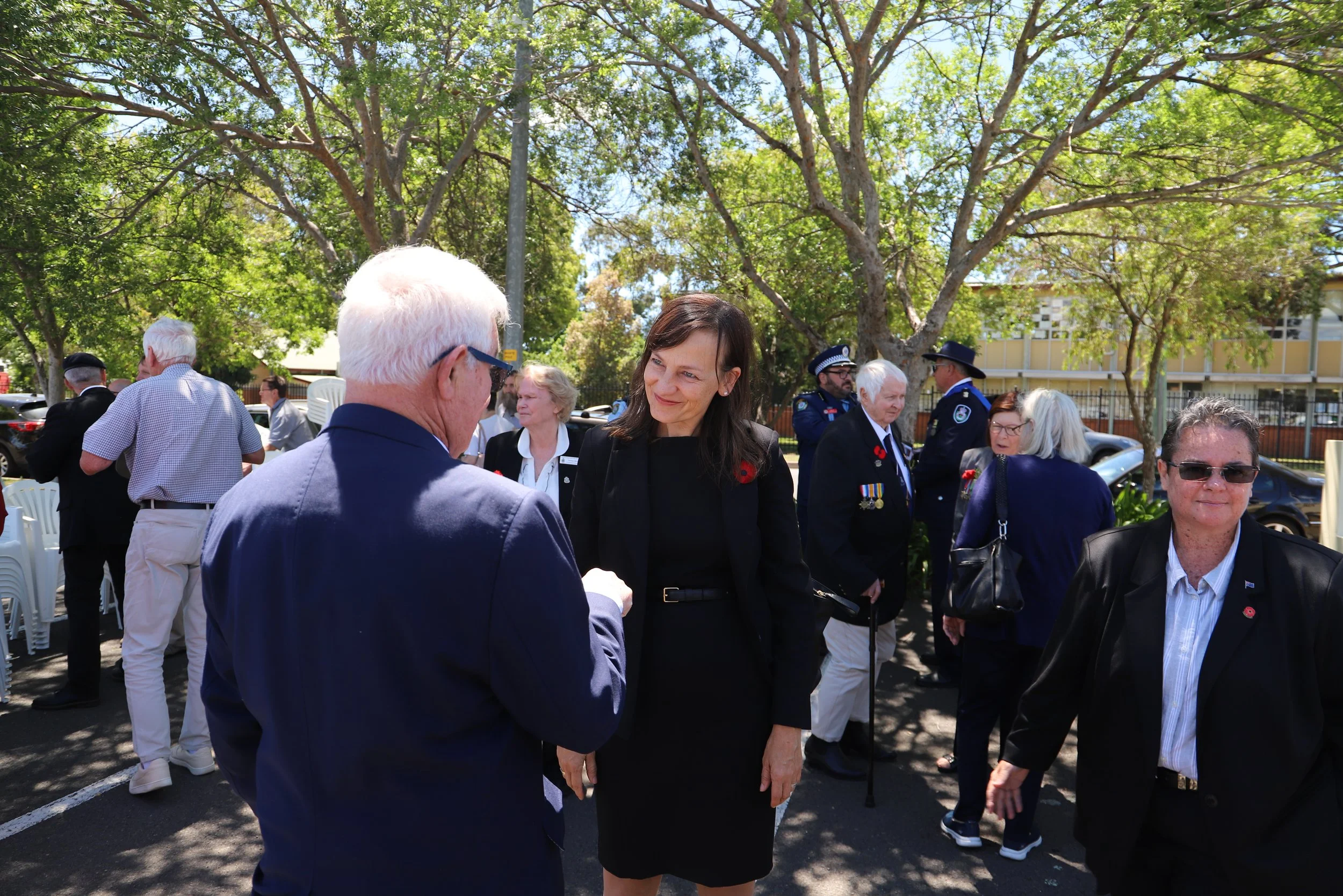 Carol Berry MP attending an outdoor Remembrance Day ceremony, some people wearing suits and medals, gathered under trees on a sunny day.