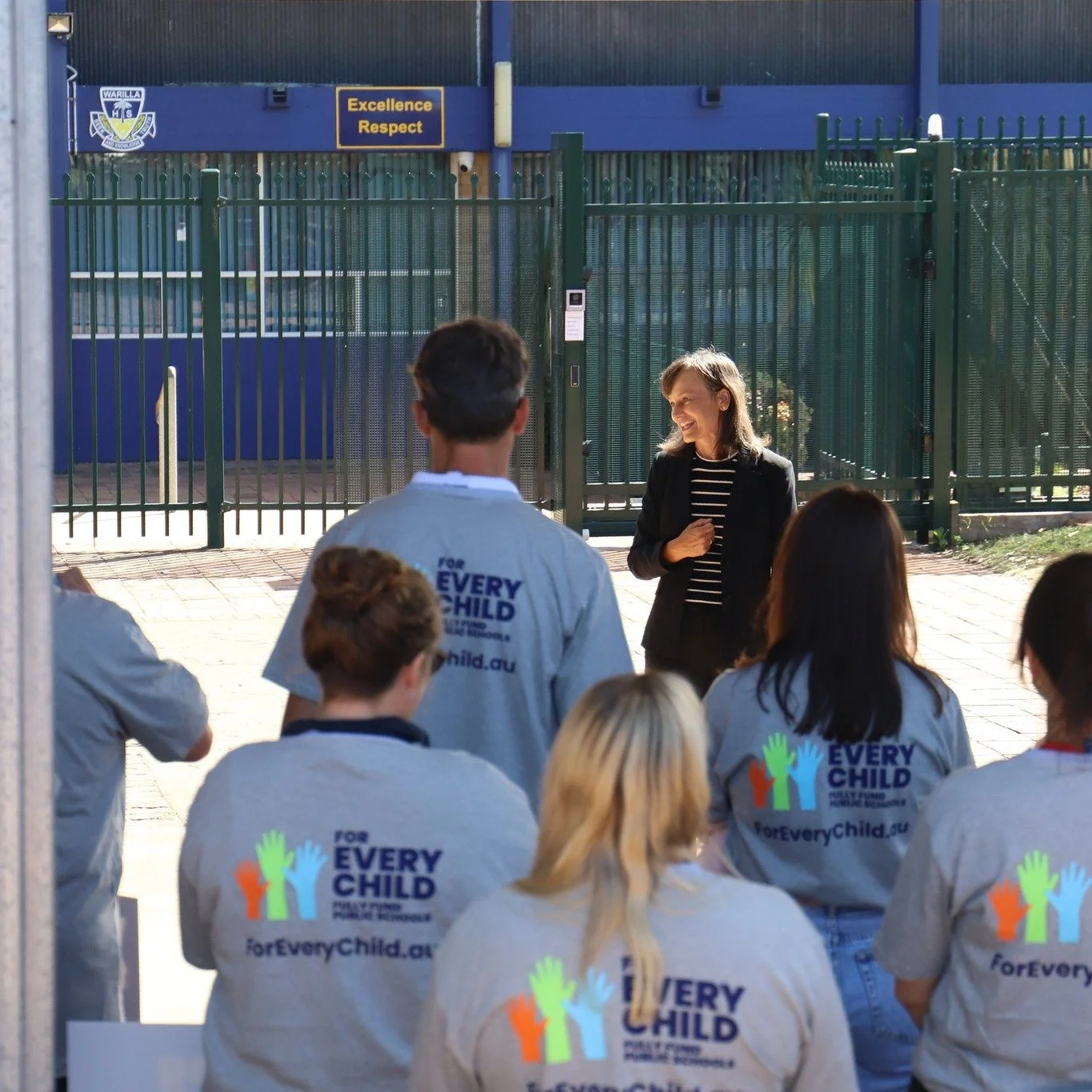A group of people wearing gray shirts with the 'Every Child' logo and website, attending a presentation or speech outdoors near a fence and a building with a sign that says 'Excellence Respect'. Carol Berry MP is speaking to the crowd.