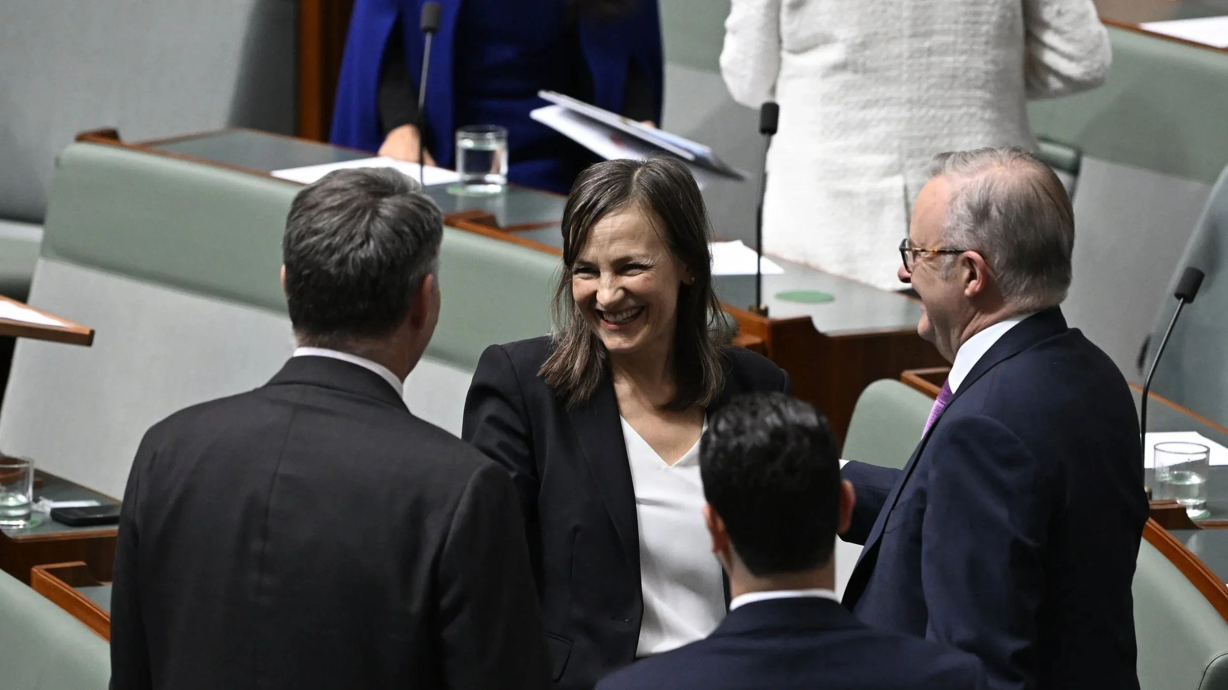 Carol Berry MP and Prime Minister Anthony Albanese, are engaged in conversation and smiling in Parliament House. The woman is at the center, wearing a black blazer and a white top, while the men are dressed in suits.
