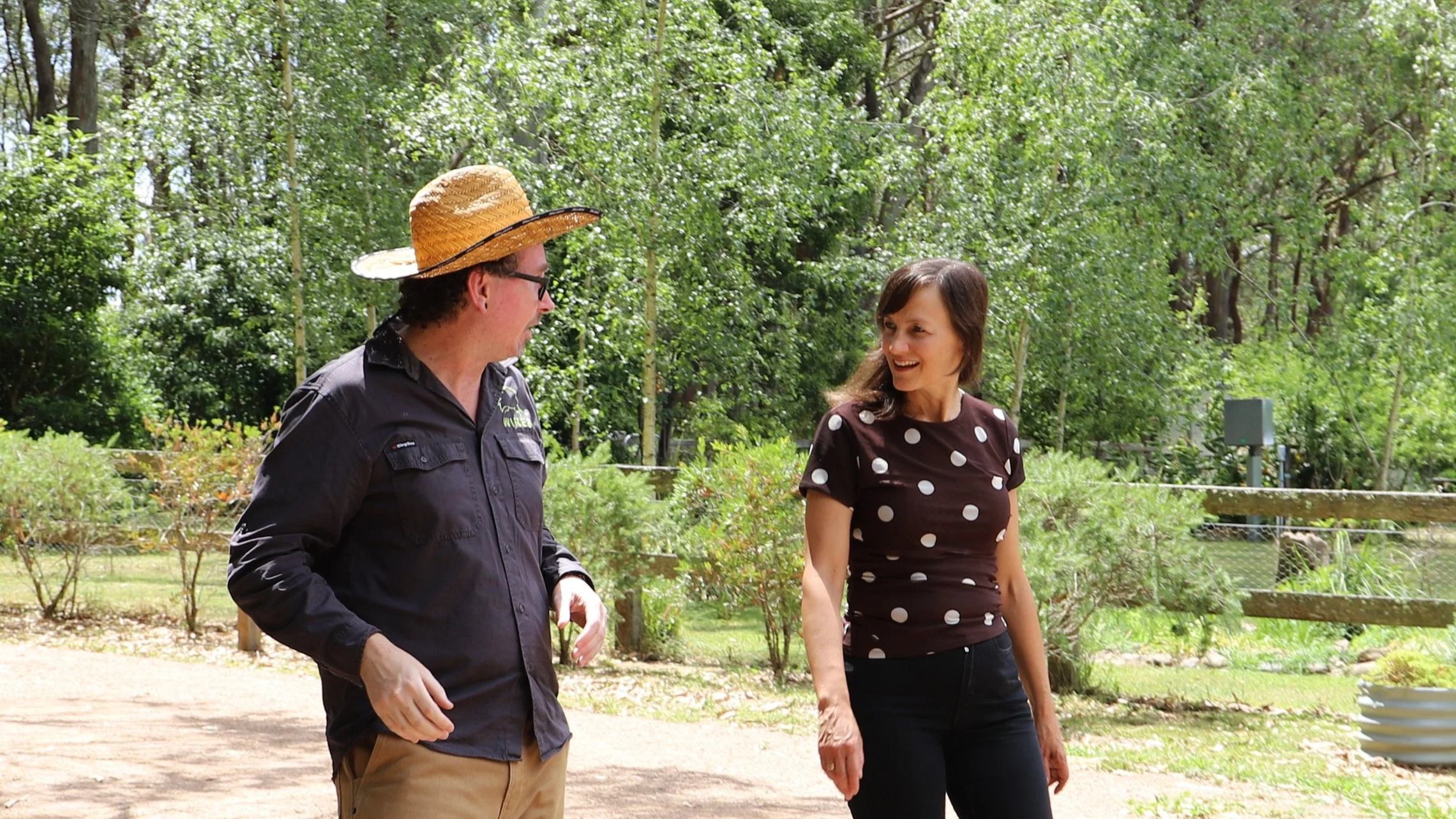A man wearing a large straw hat and black shirt with Carol Berry MP in a black shirt with white polka dots, both smiling and talking outdoors in a green, wooded area.