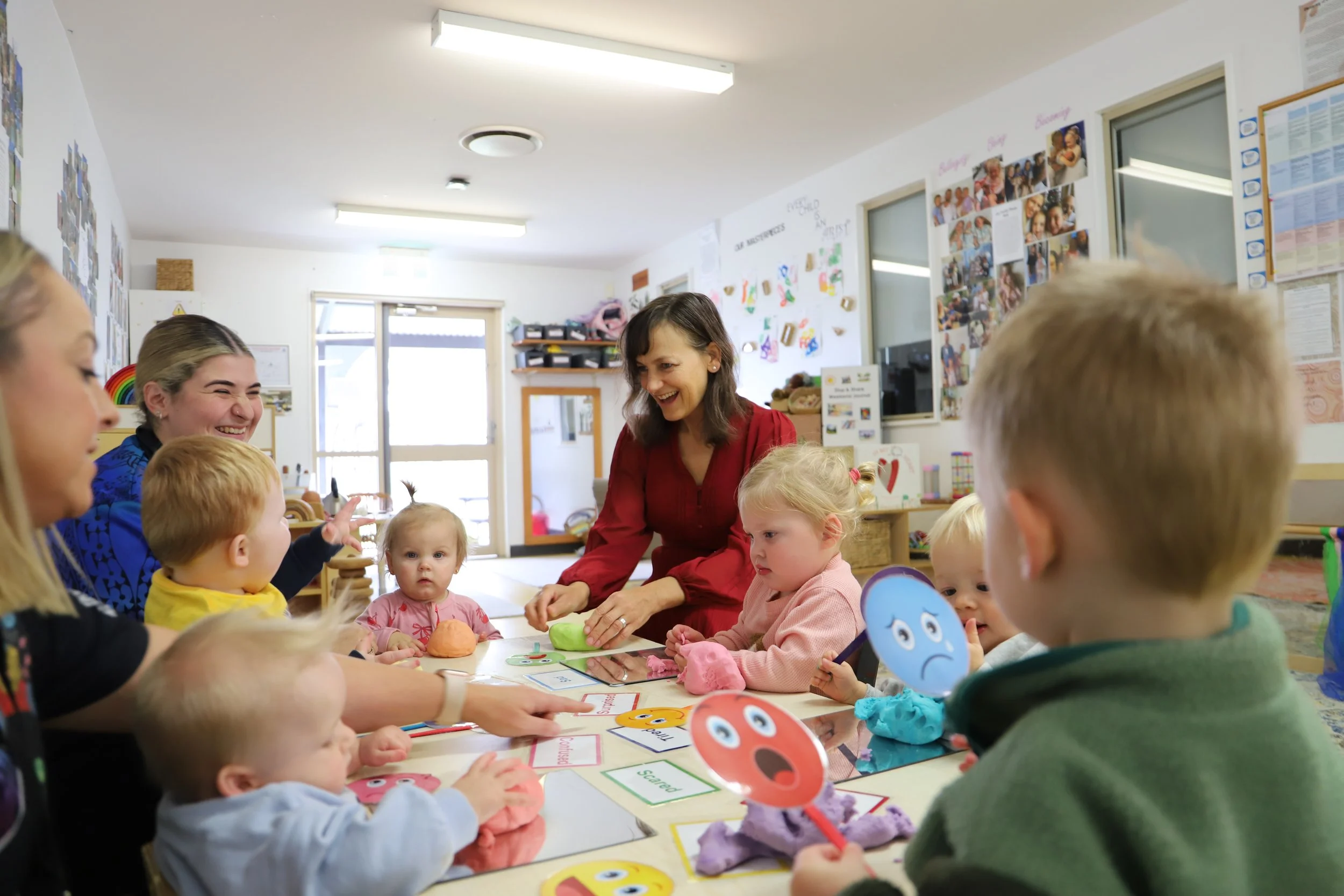 Carol Berry and children gathered at a table in a classroom, engaged in an activity, with a woman in a red shirt leading. The children are holding arts and crafts supplies and paper with emoji faces, with colorful decorations and photos on the walls.