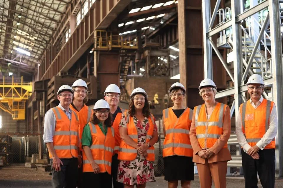 A group of eight people, including Carol Berry MP, wearing safety vests and hard hats standing inside an industrial facility.