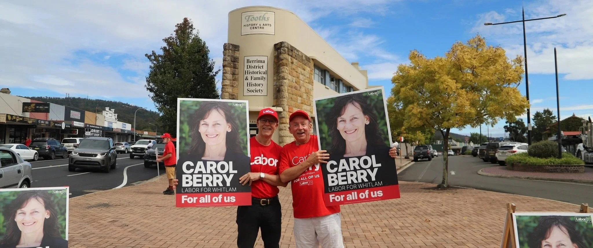 Two men standing on a sidewalk holding political campaign posters for Carol Berry, who is running for labor in Whitlam. The men are smiling, wearing red shirts and caps, and standing in front of a town center with signs for a history and arts center, trees with autumn foliage, and a street with parked and moving cars.