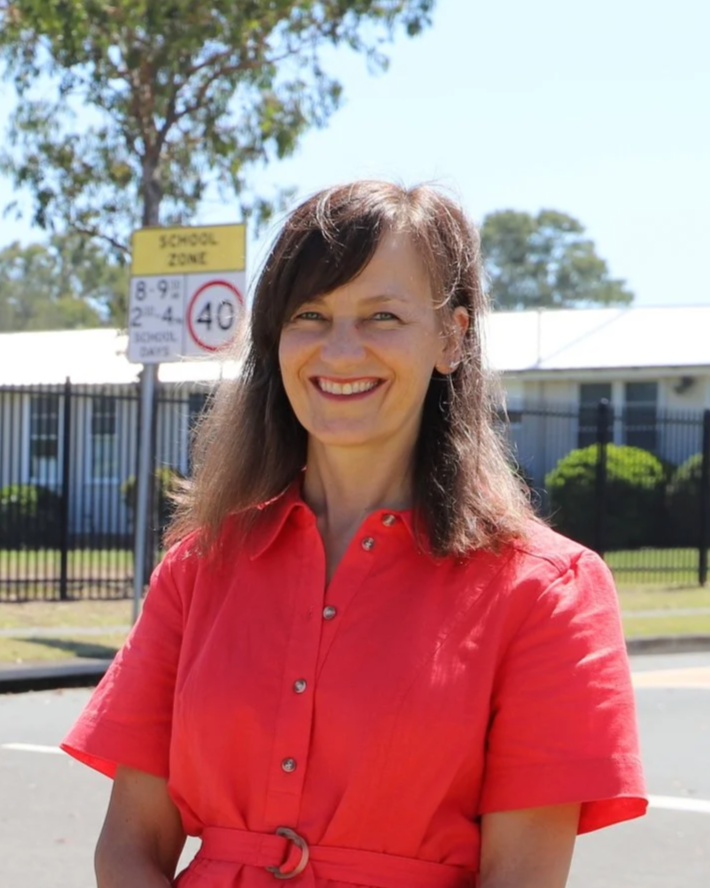 A photo of Carol Berry MP, a women with long brown hair smiling, wearing a red shirt, standing outdoors.