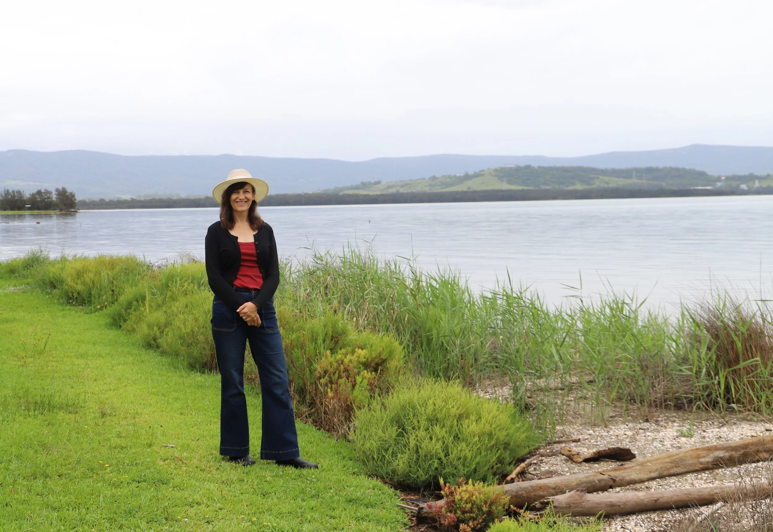 Carol Berry MP standing on grassy shoreline near a body of water, wearing a wide-brimmed hat, black cardigan, red top, and jeans, smiling with hills and cloudy sky in the background.