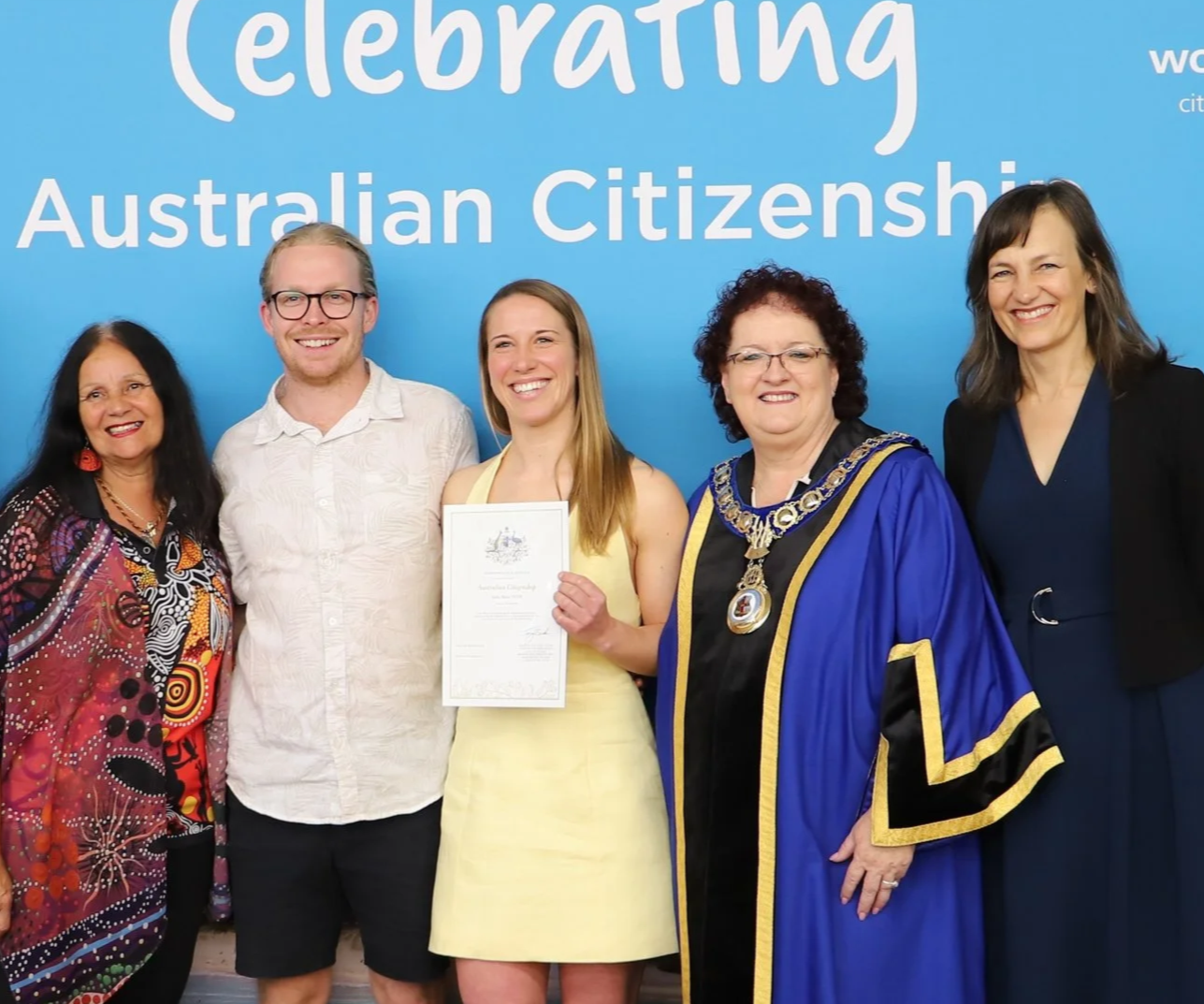 Group of five four people, and Carol Berry MP, standing in front of a blue background with the words 'Celebrating Australian Citizenship.' One woman in the center is holding a certificate and smiling.