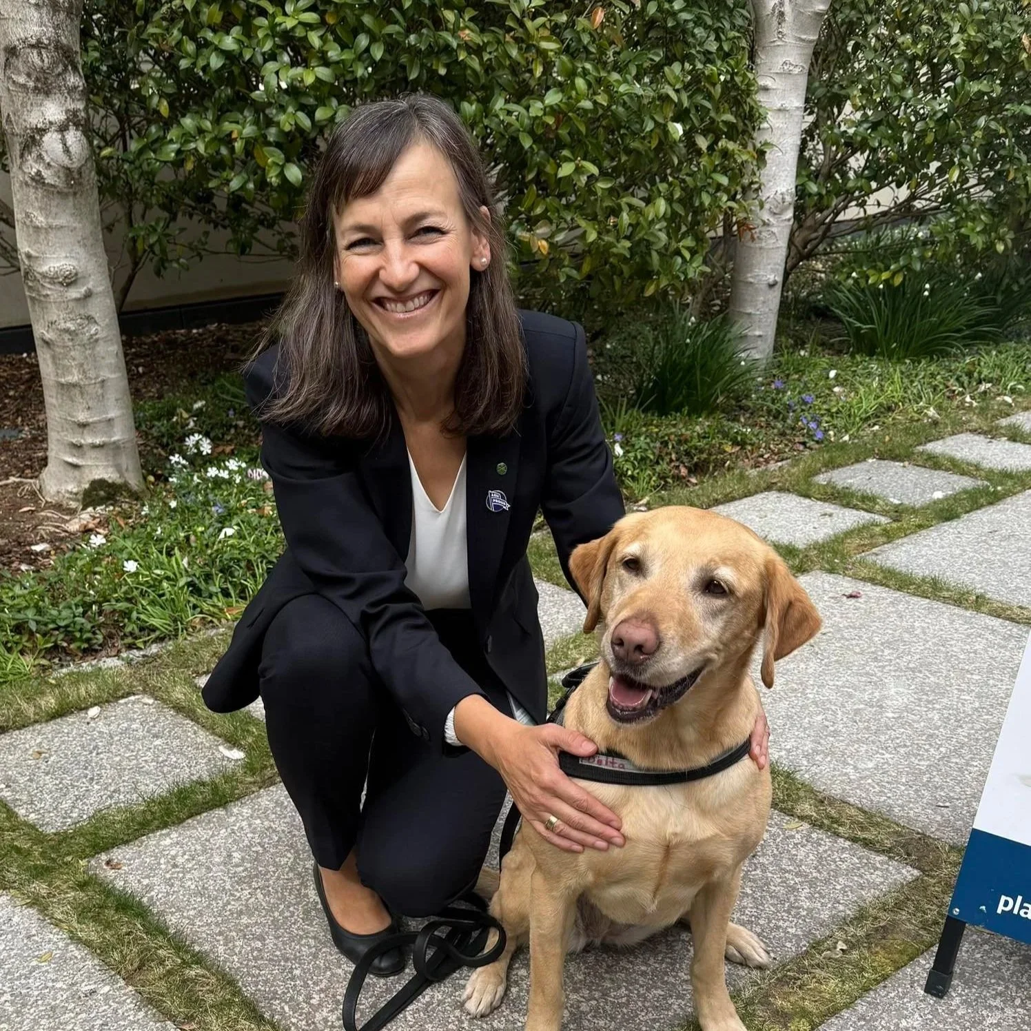 Carol Berry MP, a woman with shoulder-length brown hair, smiling, wearing a black blazer and white blouse, kneeling next to a yellow Labrador Retriever dog outdoors on a stone pathway surrounded by greenery and trees.