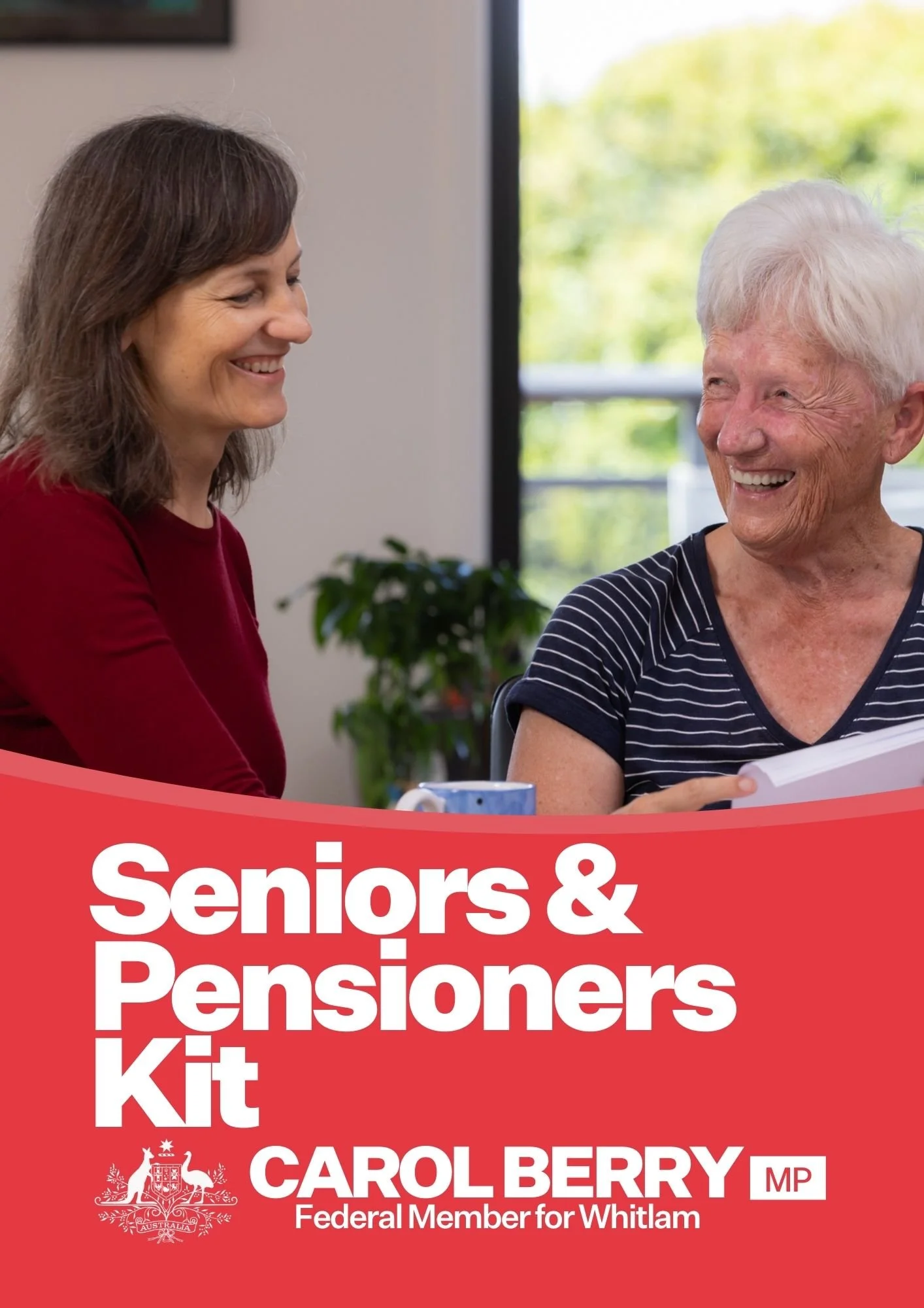 Carol Berry MP and an elderly woman smiling and talking in a room with a large window and a plant in the background. The elderly woman is holding papers, and there's a mug on the table.