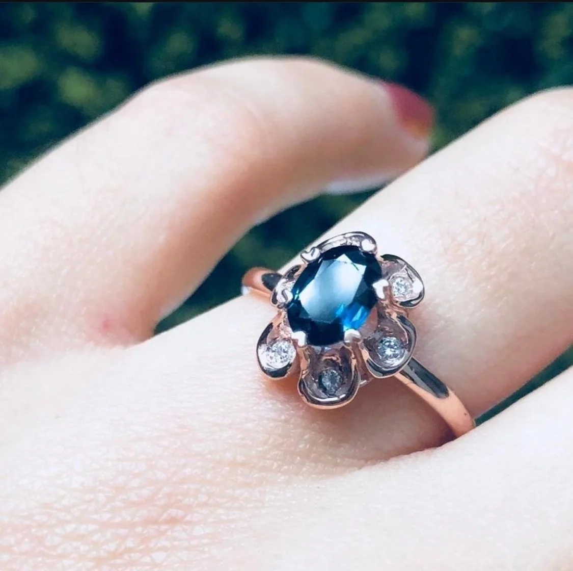 Anillo de plata con piedra azul central y detalles de diamantes en una mano con fondo desenfocado.