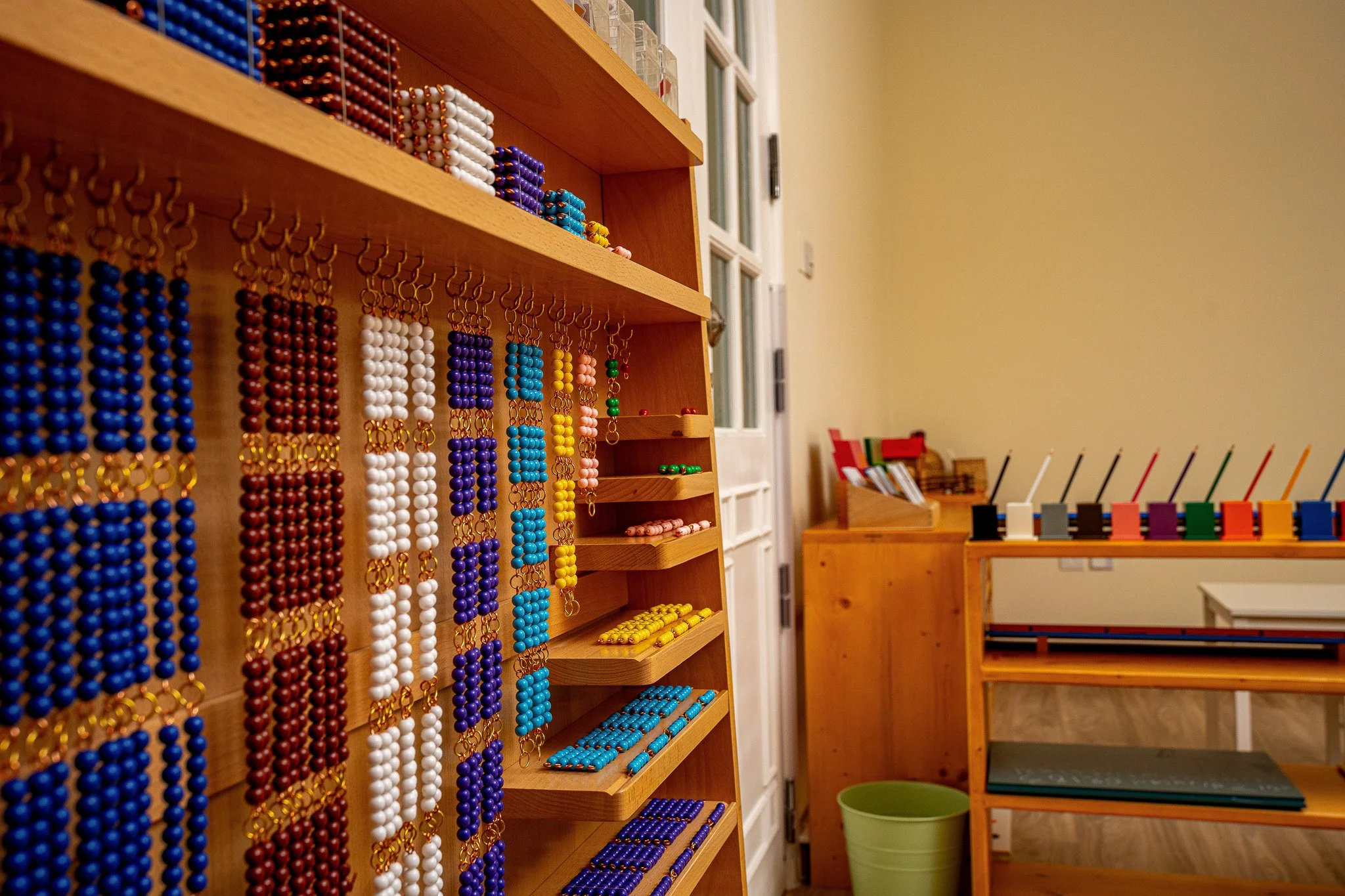 عرض مجوهرات ملونة من الخرز معلقة على الرفوف في فصل دراسي أو غرفة حرفية مع أثاث خشبي وجدار فاتح اللون.