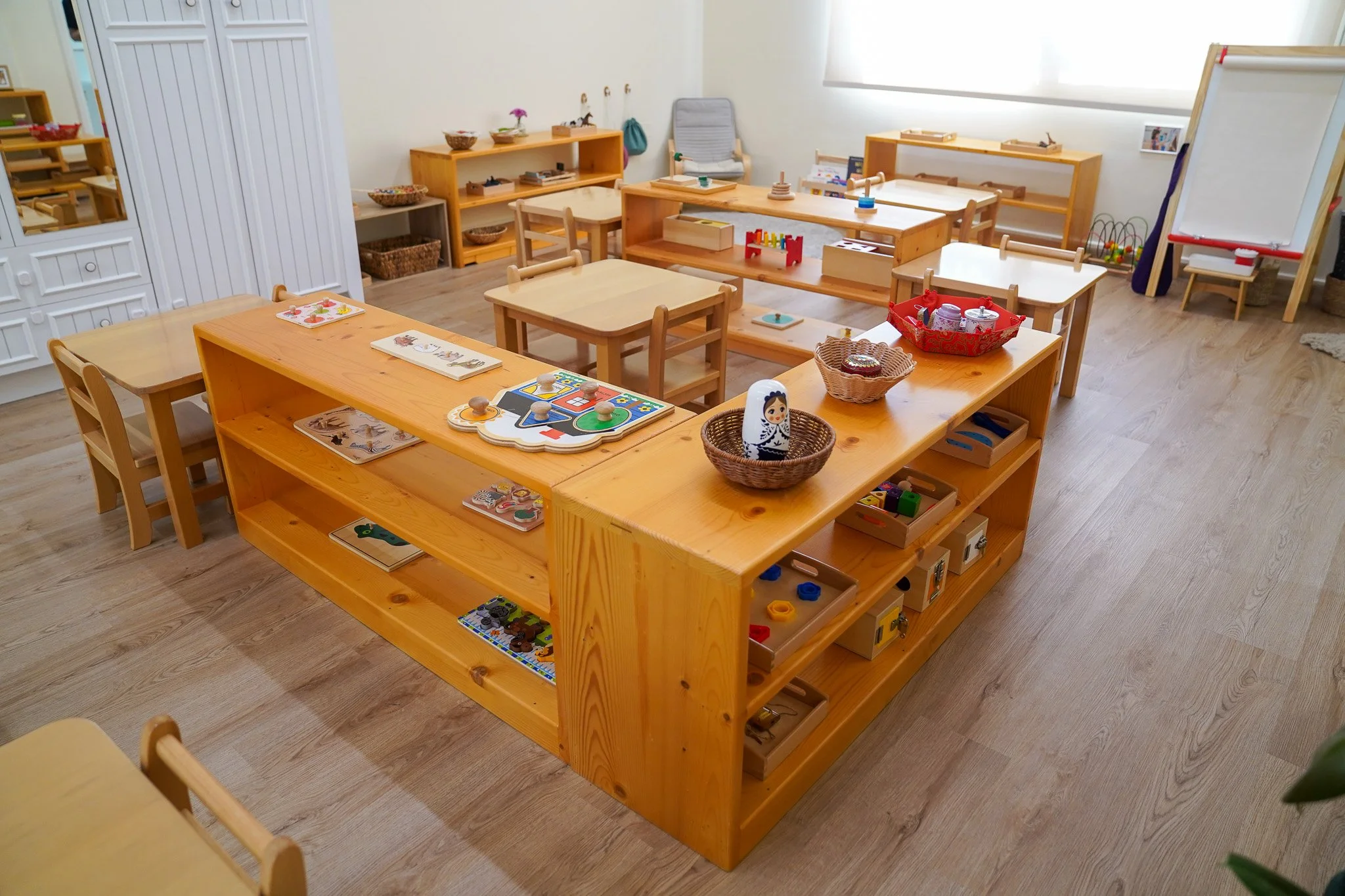 فصل مونتسوري فارغ يحتوي على طاولات وكراسي خشبية ورفوف مع ألعاب وألعاب تعليمية وسبورة بيضاء.