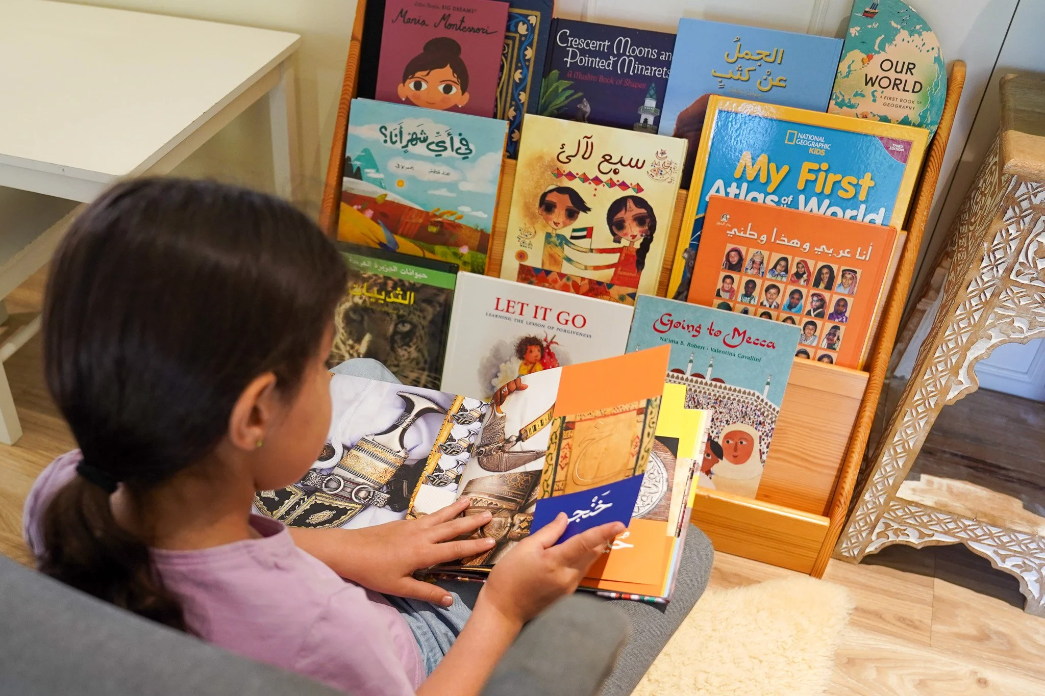 فتاة صغيرة بشعر داكن مربوط إلى الخلف، تجلس وتقرأ كتابًا للأطفال في غرفة ذات أرضية خشبية. أمامها رف كتب خشبي أو حامل مجلات خشبي مليء بكتب الأطفال بلغات ومواضيع مختلفة، بما في ذلك الإسلام والجغرافيا والتعليم المبكر. بعض الكتب معروضة وأغلفتها متجهة إلى الأمام، ويبدو أنها تقرأ أو تتفحص الكتاب المفتوح الذي تحمله.