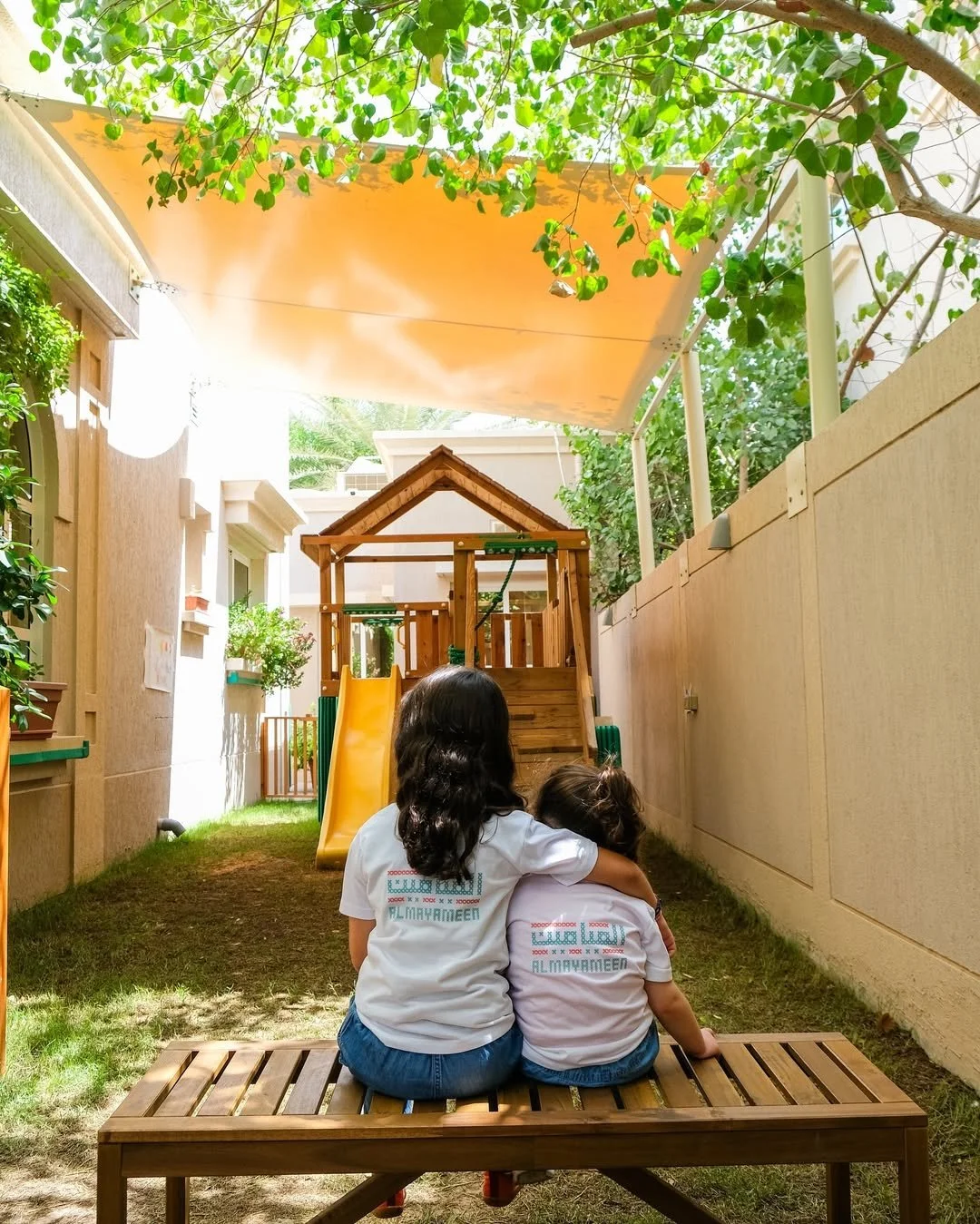 طفلان يجلسان على مقعد خشبي في فناء خلفي في مواجهة الكاميرا، وفي الخلفية منزل خشبي للعب وزلاجة مظللة بأوراق الشجر ومظلة برتقالية اللون.