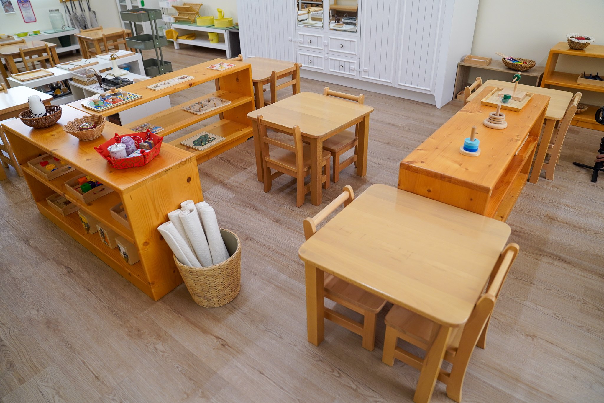 فصل دراسي أو غرفة ألعاب بها عدة طاولات وكراسي خشبية ورفوف بها سلال وألعاب وبعض المواد التعليمية.