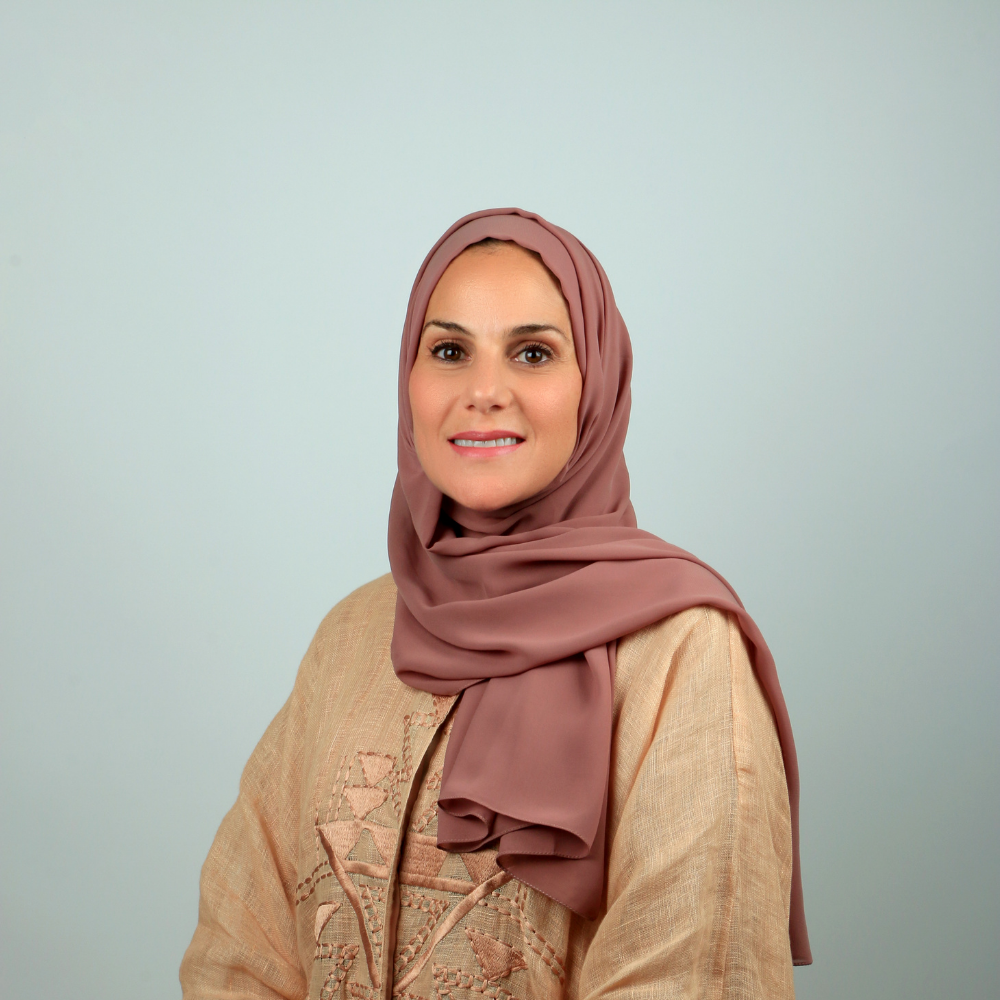 صورة لامرأة مبتسمة ترتدي قميصاً مطرزاً باللون البيج وغطاء رأس بنفسجي على خلفية زرقاء فاتحة.