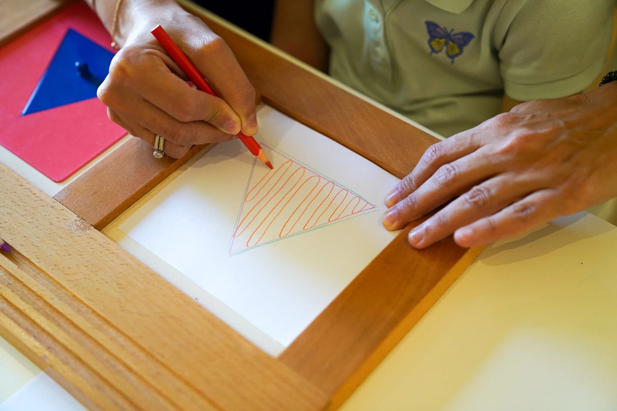 شخص يرسم مثلثاً بحدود ملونة وخطوط برتقالية في الداخل على ورقة، باستخدام قلم تلوين أحمر، مع أشكال هندسية على طاولة.