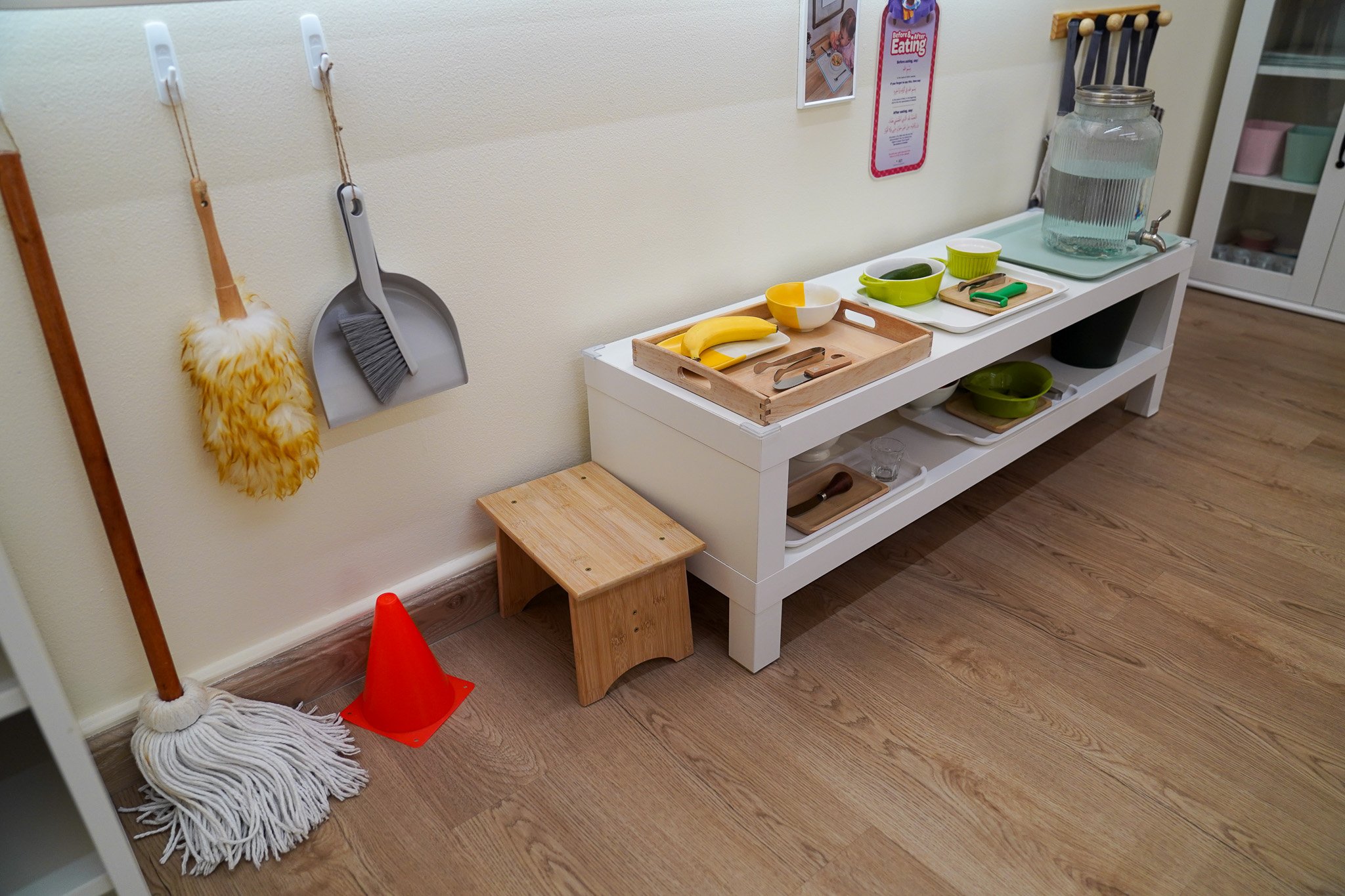 منطقة لعب للأطفال تم إعدادها لتبدو وكأنها مطبخ مع أطباق ألعاب وفاكهة ولوازم تنظيف مع كرسي خشبي صغير وأدوات تنظيف في غرفة ذات أرضية خشبية.