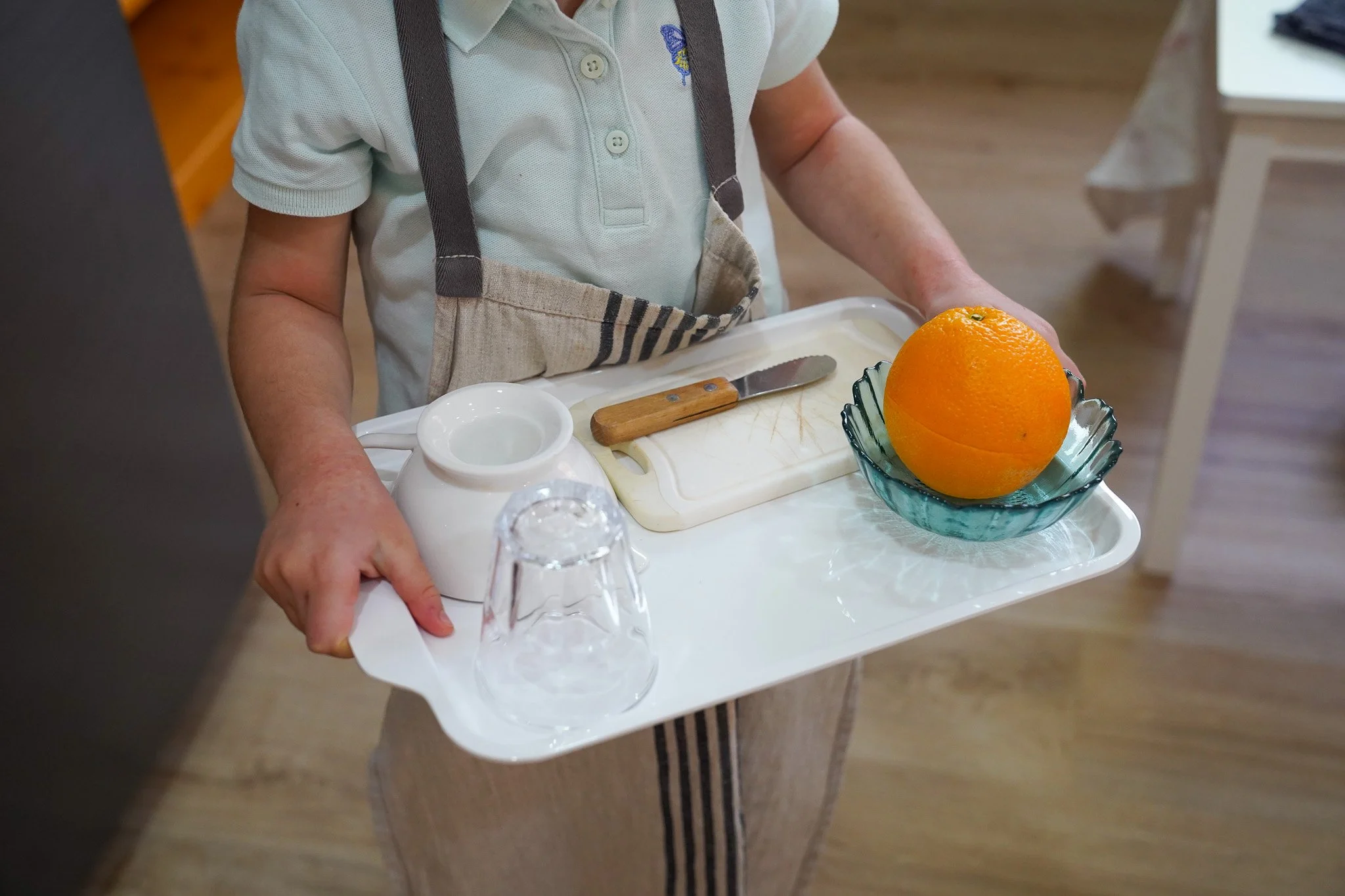 طفل يرتدي مئزرًا مخططًا ويحمل صينية بها كأس وكوب أبيض ولوح تقطيع وسكين ووعاء برتقال.