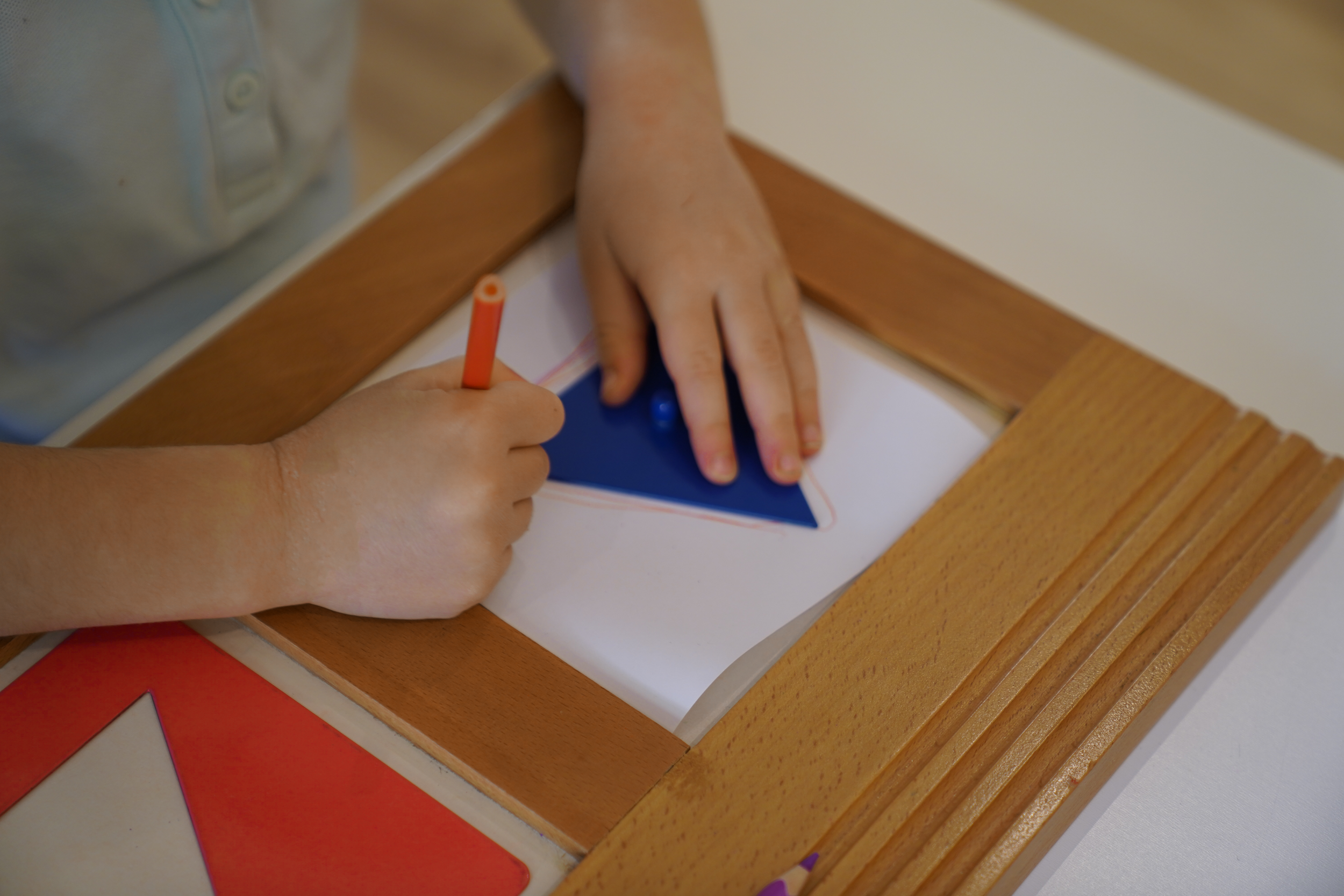 يدا طفل تلونان مثلثاً ورقياً بقلم تحديد أحمر على لوحة رسم خشبية.