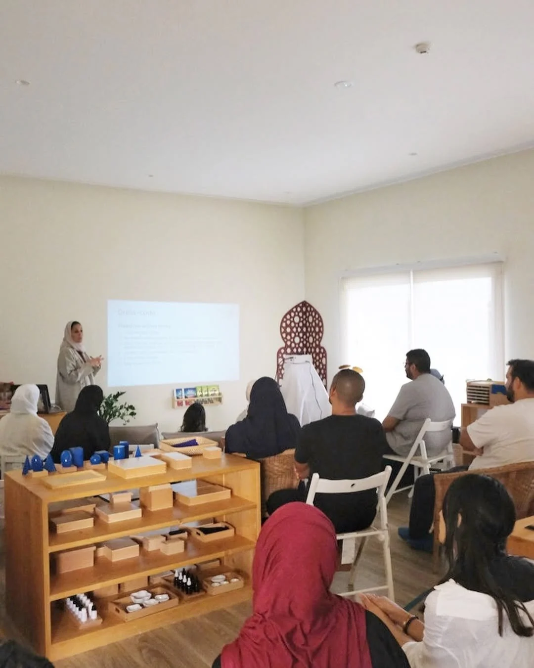 امرأة تقدم عرضًا تقديميًا لمجموعة من الأشخاص المتنوعين في غرفة مضاءة جيدًا ذات نوافذ كبيرة. يوجد رف خشبي به مواد تعليمية ولوحة خشبية مزخرفة في الخلفية.